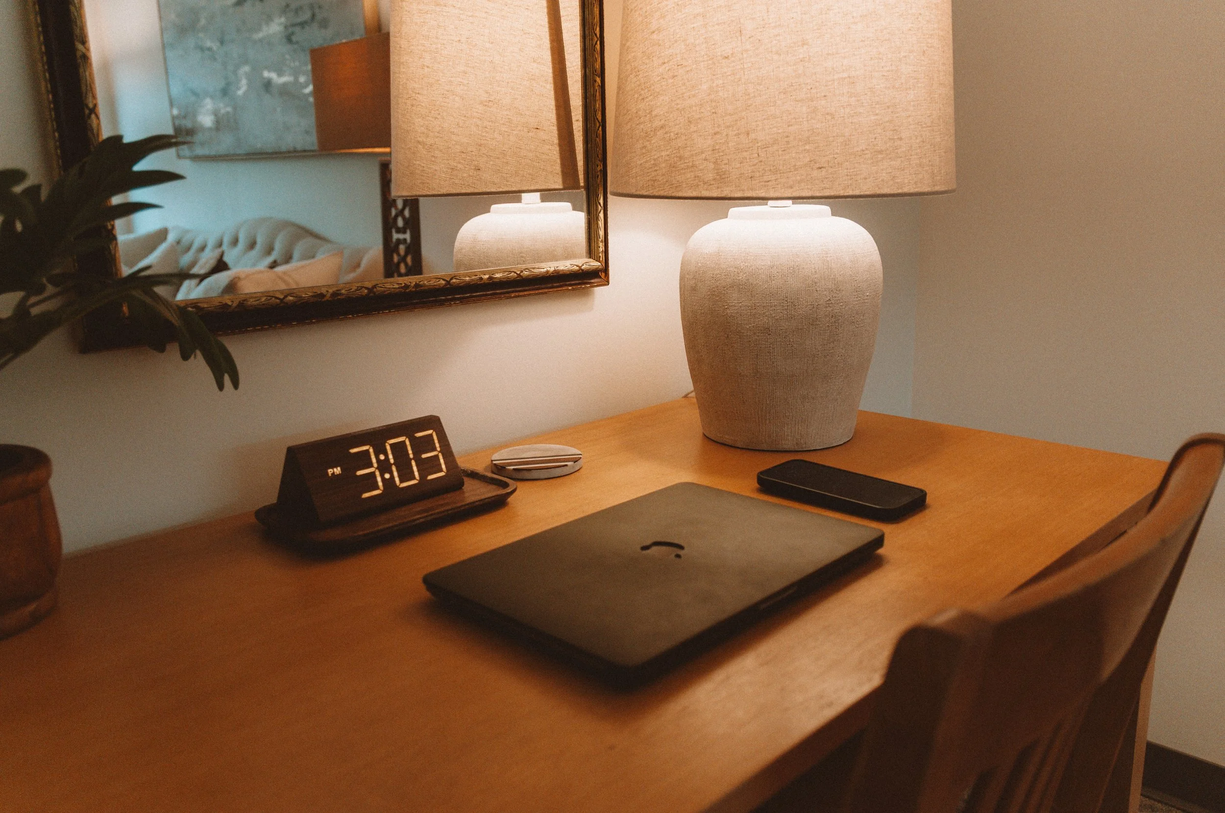 Wooden desk with a digital clock showing 3:03 PM, a closed laptop, a smartphone, a small round device, a table lamp, and a mirror reflecting part of a bed with a tufted headboard.