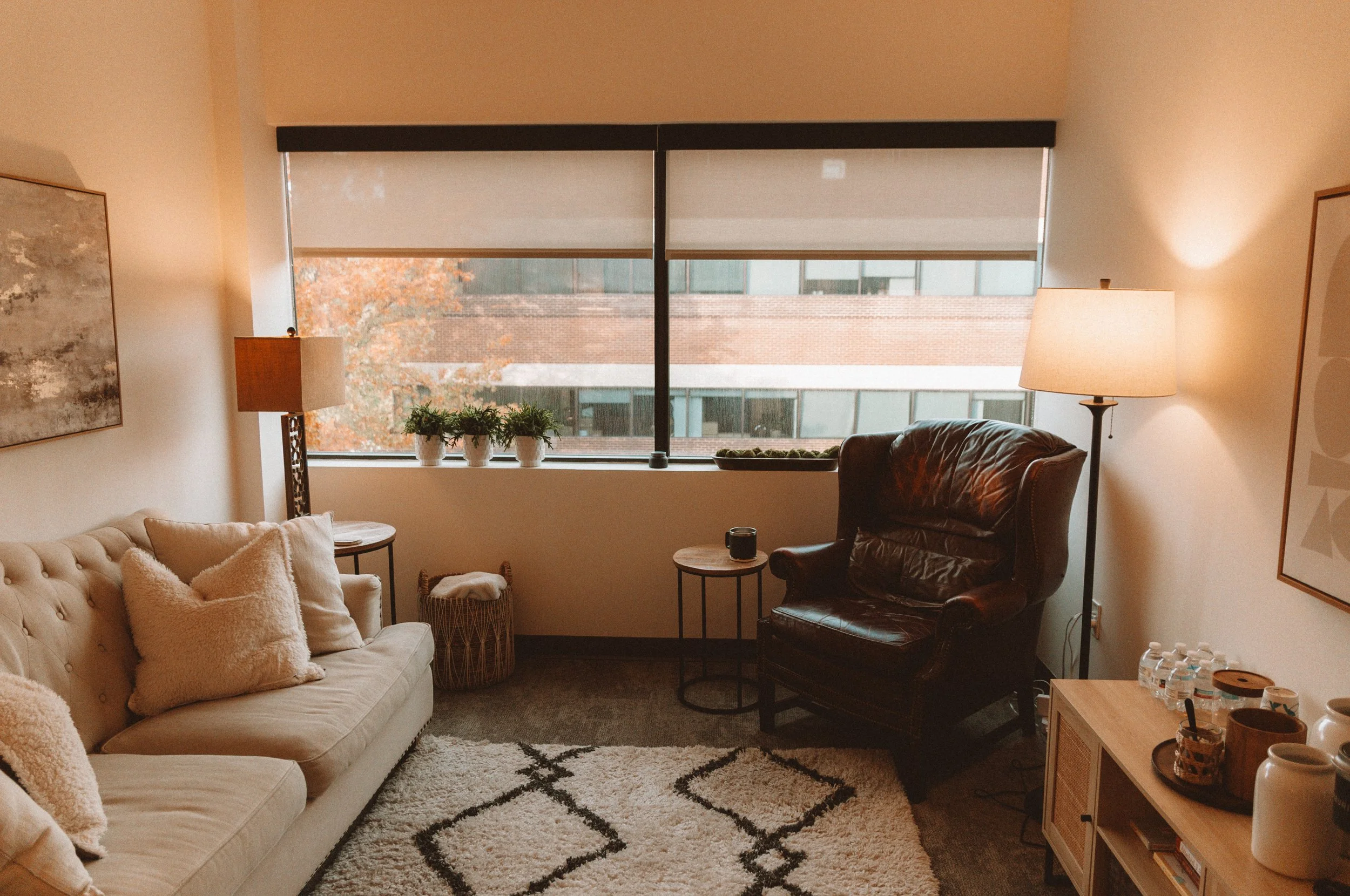 Living room with beige sofa, decorative pillows, leather armchair, side tables, table lamps, potted plants on window sill, framed artwork, window with view of brick building and trees outside, and a wooden console with water bottles and decor items.