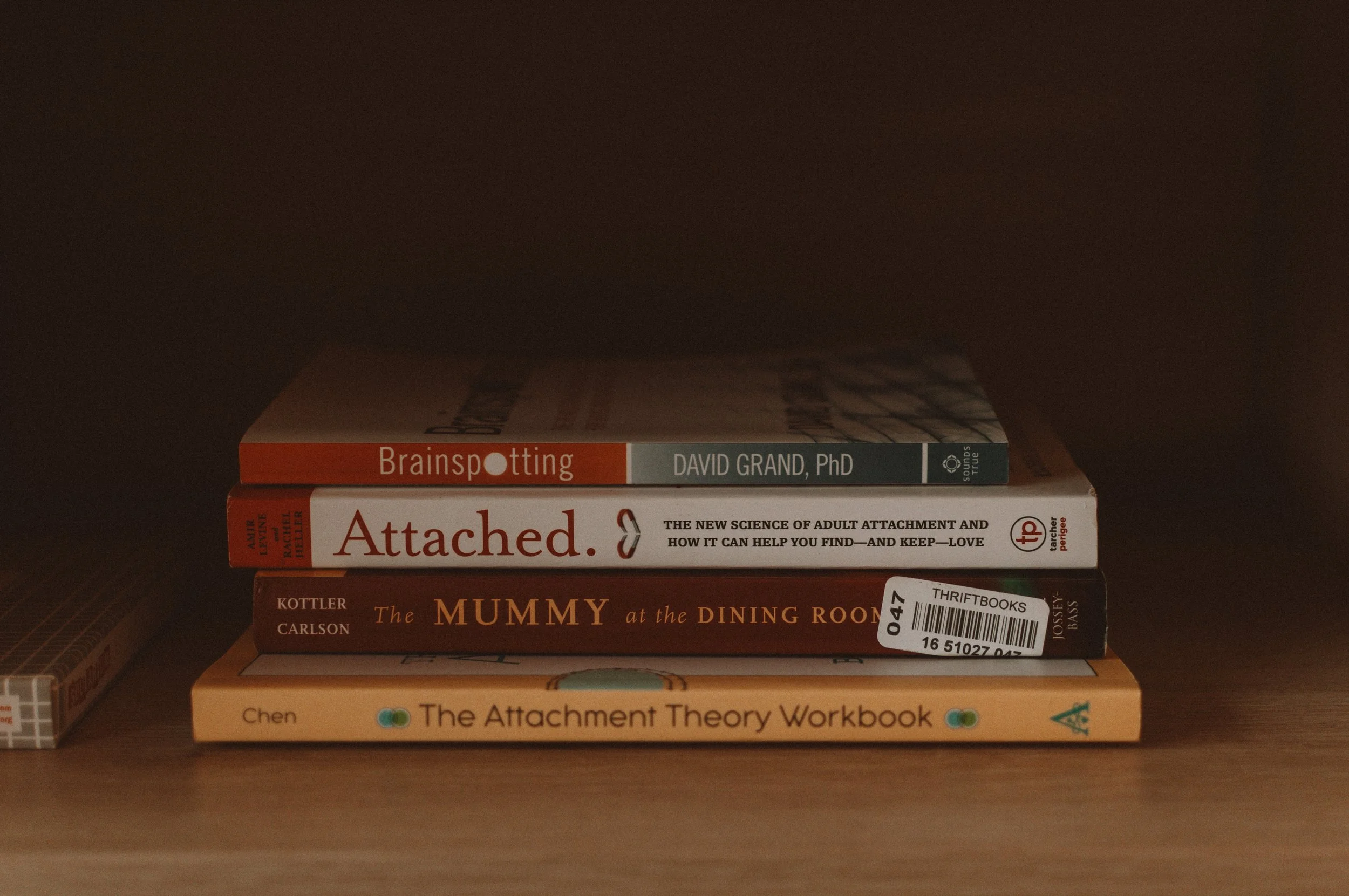 Stack of four books on a wooden surface, with titles visible: "Brainspotting" by David Grand, PhD; "Attached"; "The Mummy at the Dining Room" by Kottler Carlson; and "The Attachment Theory Workbook" by Chen.