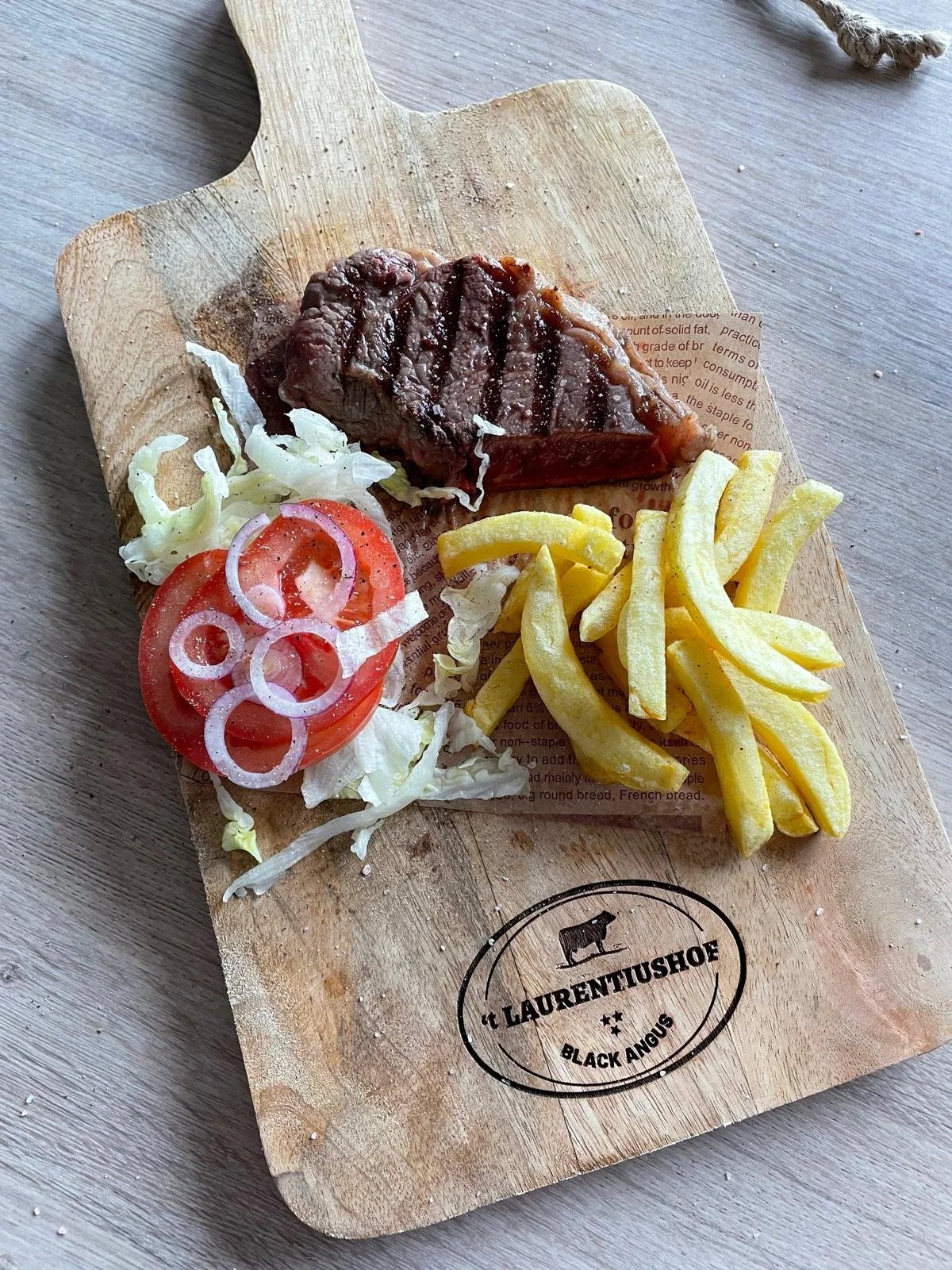 Gevormde plak met een stuk rundvlees, salade, tomaat met uienringen en frieten, op een houten snijplank met het logo 't LAURENTUSHOF' en 'BLACK ANGUS'.