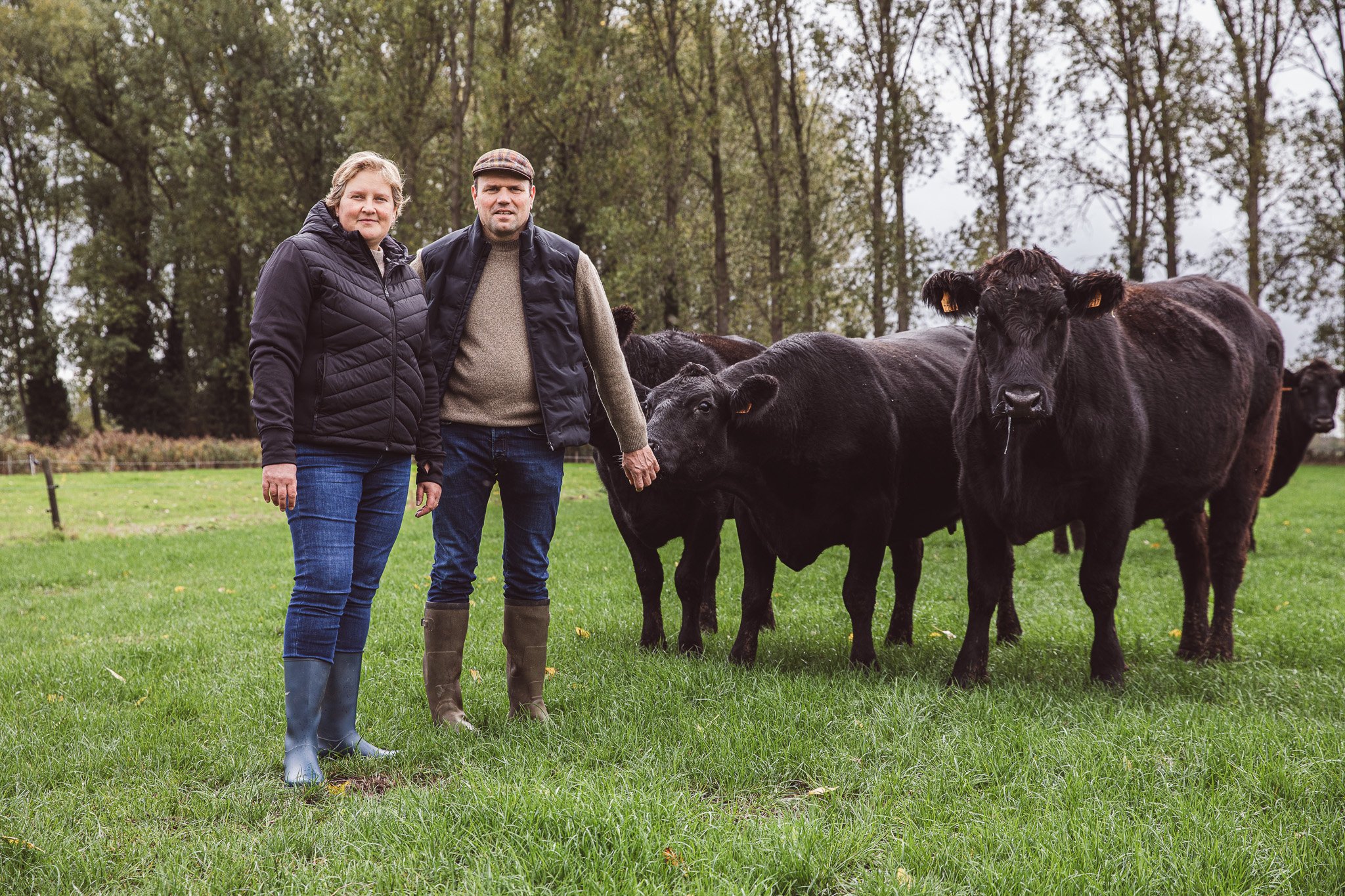 Een man en een vrouw staan op een grasveld met jonge kalfjes en koeien, bomen op de achtergrond, herfstachtig weer.