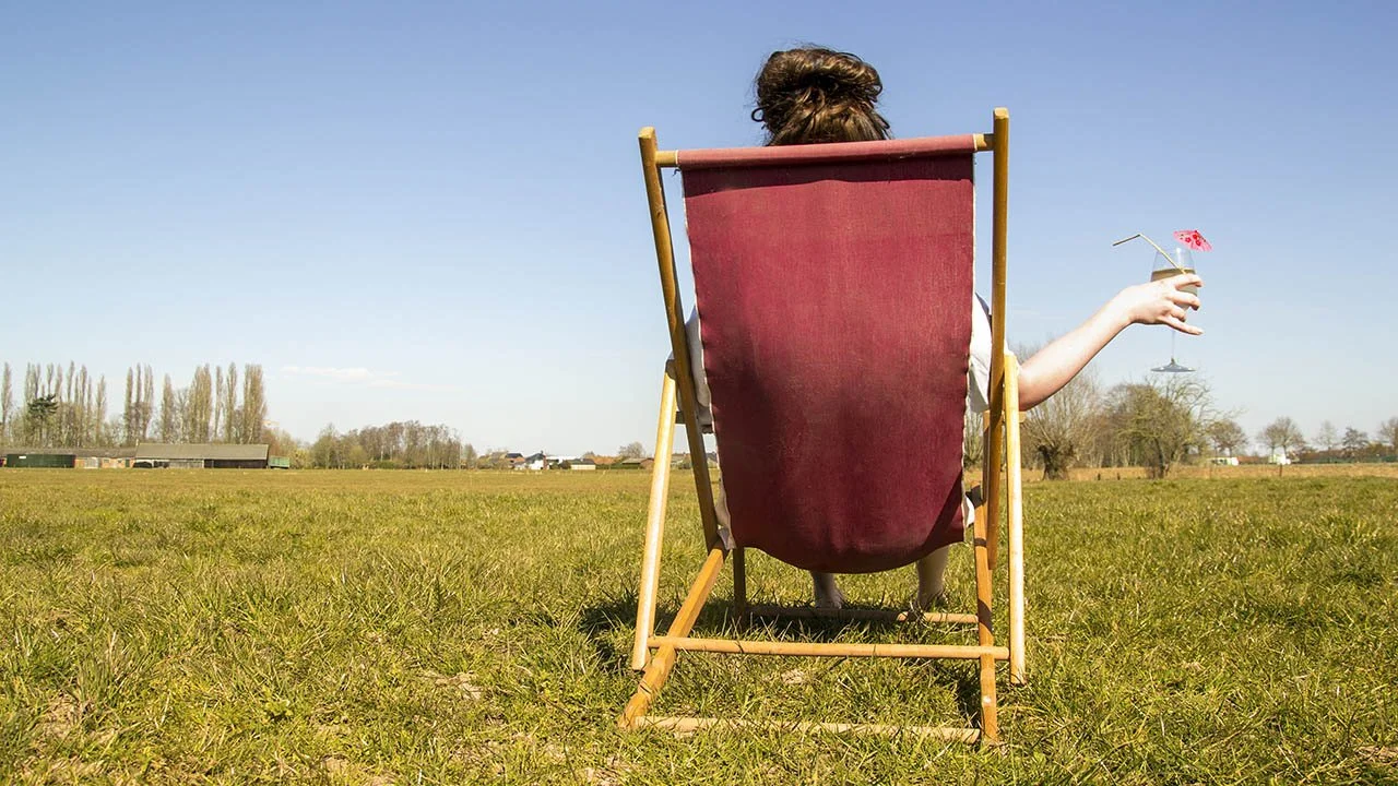 Een persoon zit in een ligstoel op een open veld, met een cocktail in de hand en een paraplu erin, onder een heldere blauwe lucht.