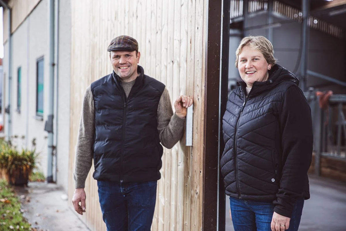 Een man en een vrouw staan buiten, leunend tegen een houten wand, en lachen naar de camera.