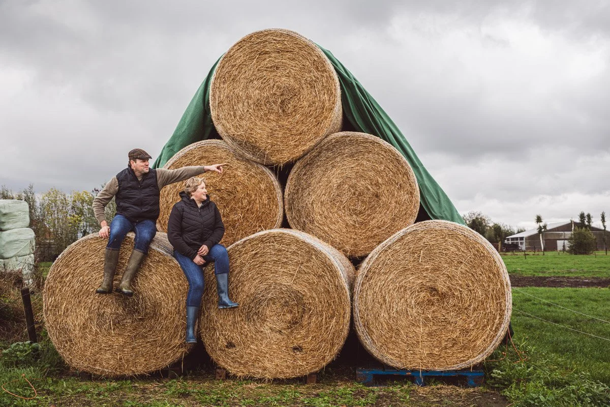 Twee mensen zitten op grote hooibalen in een open veld onder een bewolkte hemel.