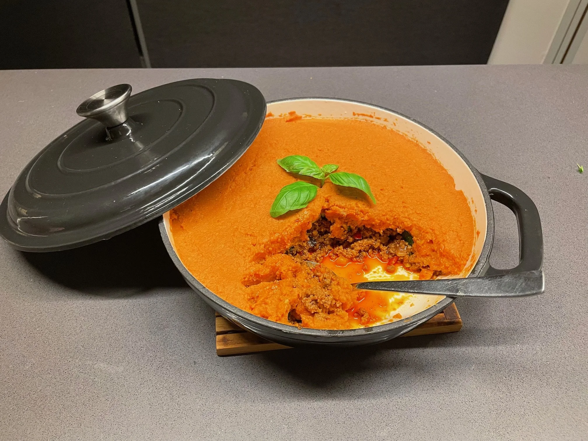 Een ovenschotel met een laag oranje zoete aardappelpuree, een vulling van gehakt en groenten, en vers geplukte basilicumblaadjes bovenop, in een zwarte ovenschaal met deksel op de rand.