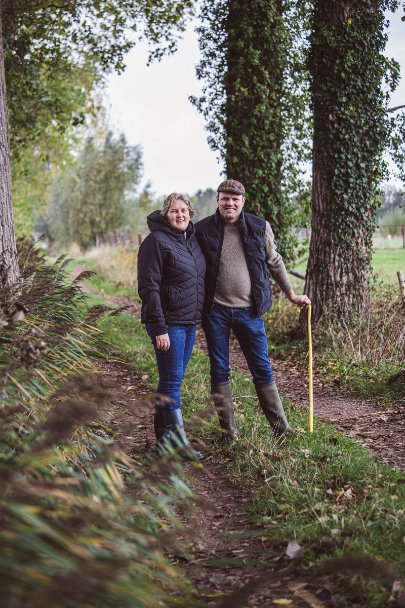 Een man en vrouw staan op een bospad, omringd door bomen, beiden dragen waterdichte kleding en laarzen.