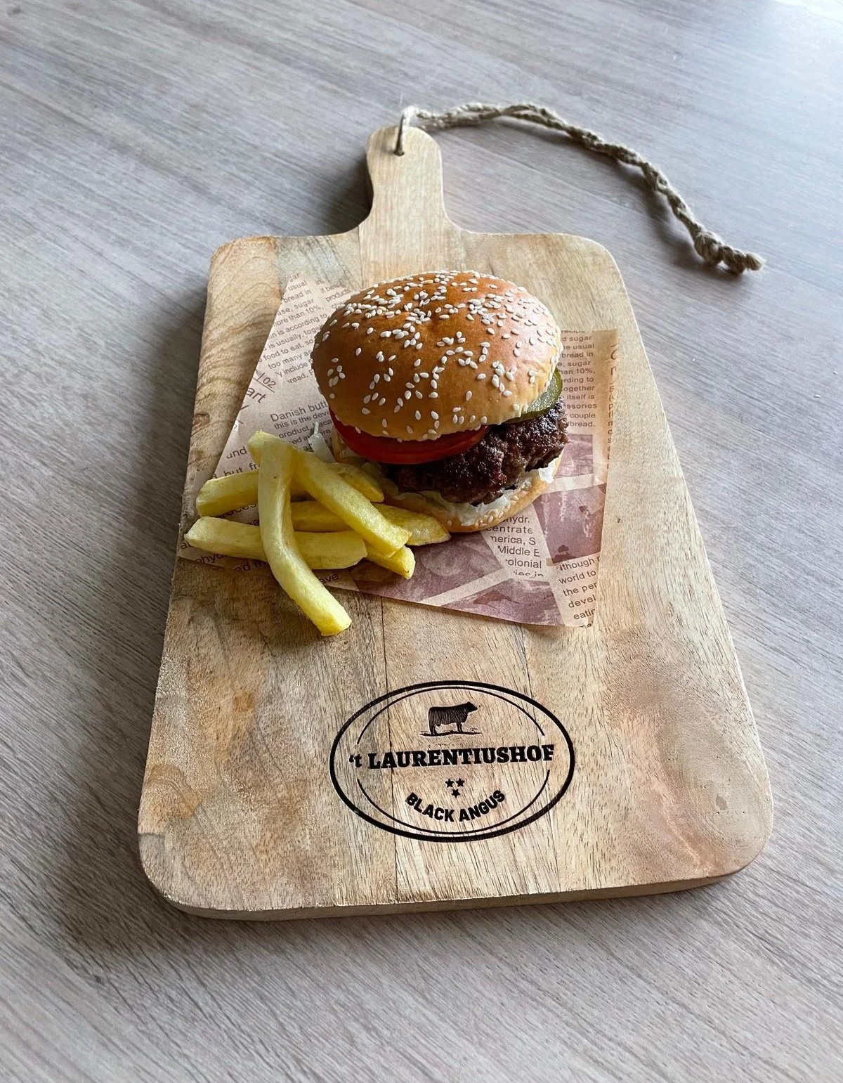 Hamburger met sesamzaadjes, tomaat en hamburgerpatty, geserveerd met Franse frieten op een houten serveerplank met Tekst 't Laurentiushof Black Angus'.