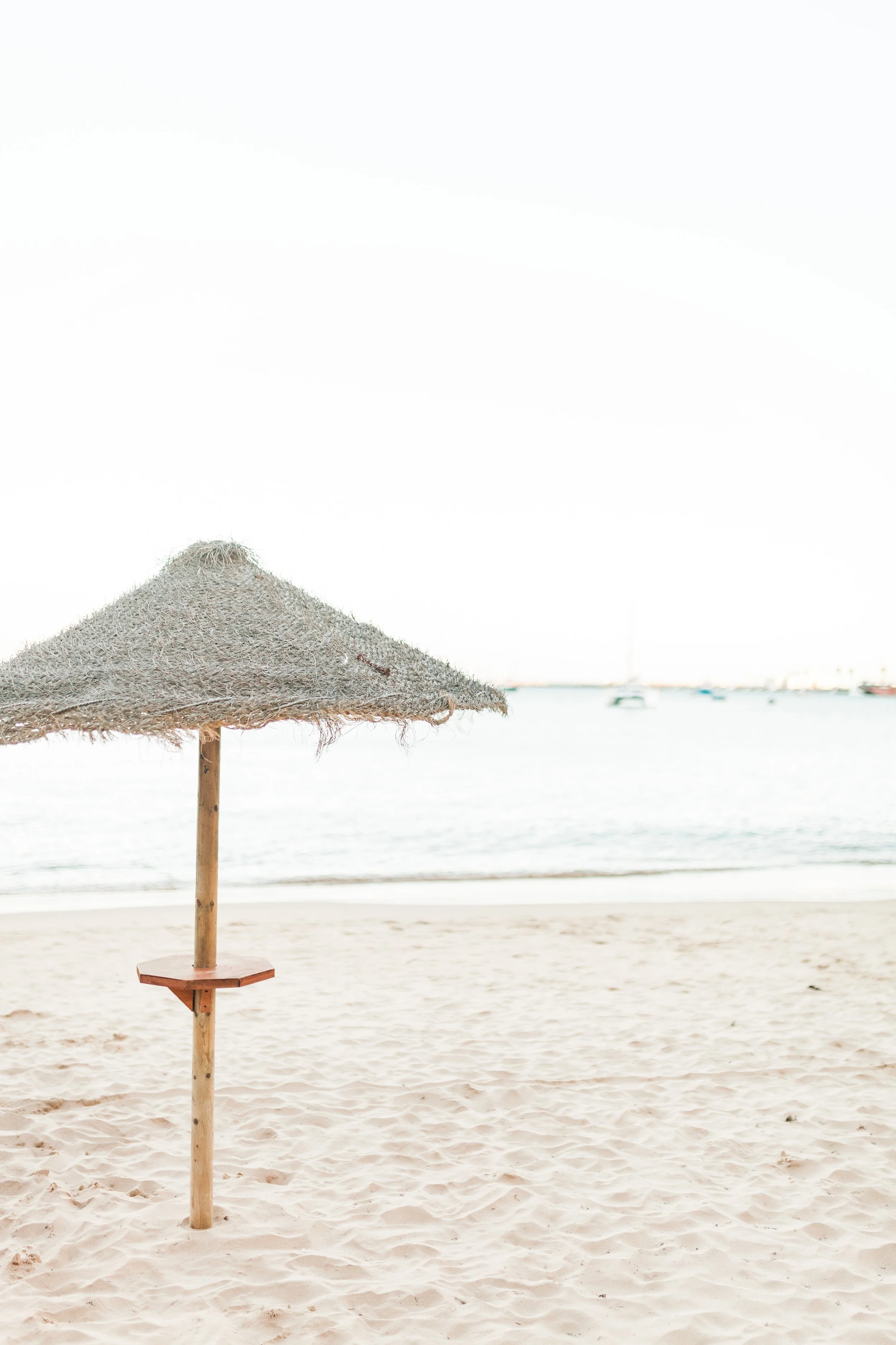 Beach scene with a sandy shore, a thatched umbrella, and boats on calm water in the background, under a clear sky.