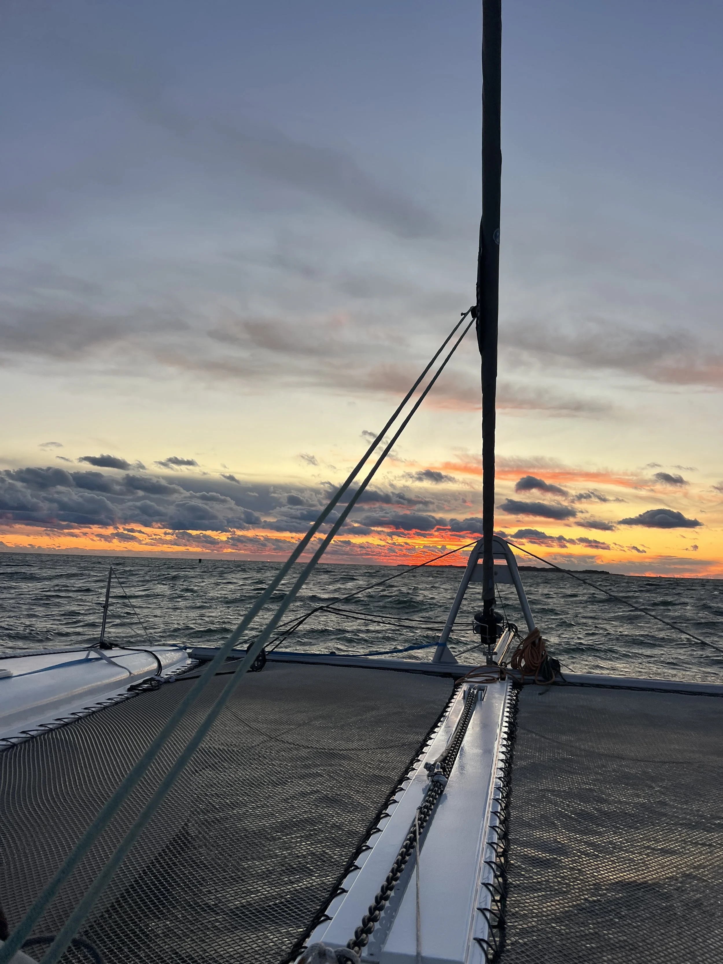 View of a boat's bow facing the ocean during sunset, with colorful clouds and the horizon in the background.