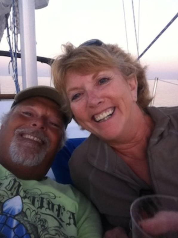 A man and woman smiling for a selfie on a boat at sunset, with water and sky in the background.