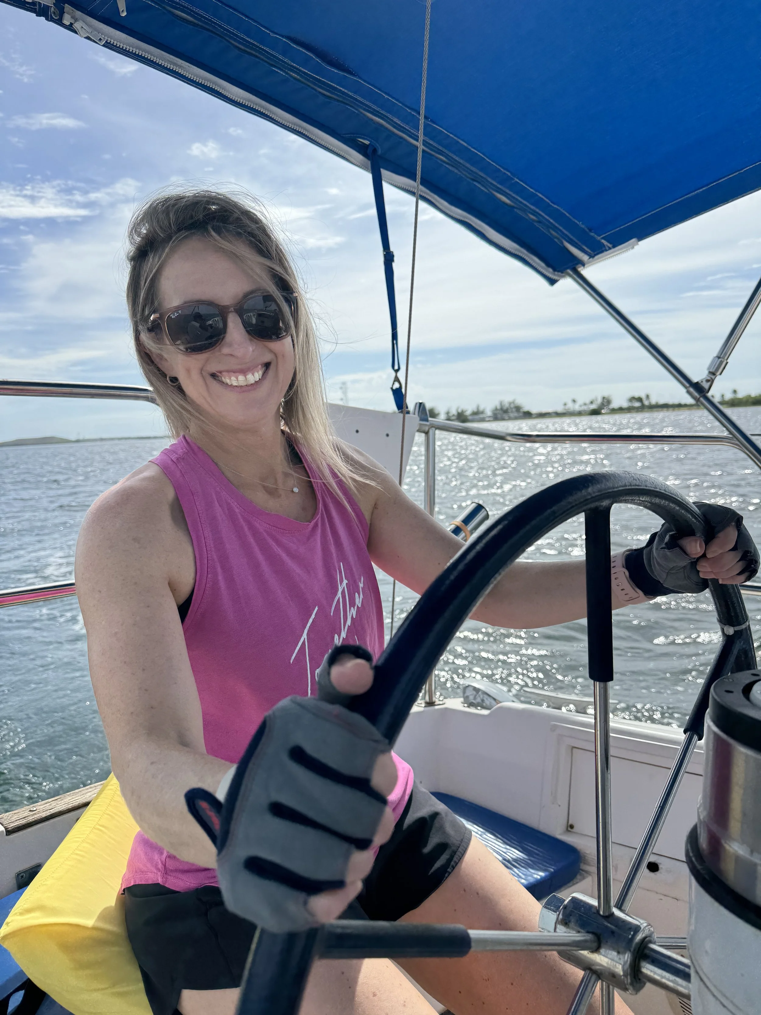 A woman with blonde hair, wearing sunglasses, a pink sleeveless top, and black shorts, smiling while steering a sailboat on a sunny day with calm water and a blue sky with clouds.