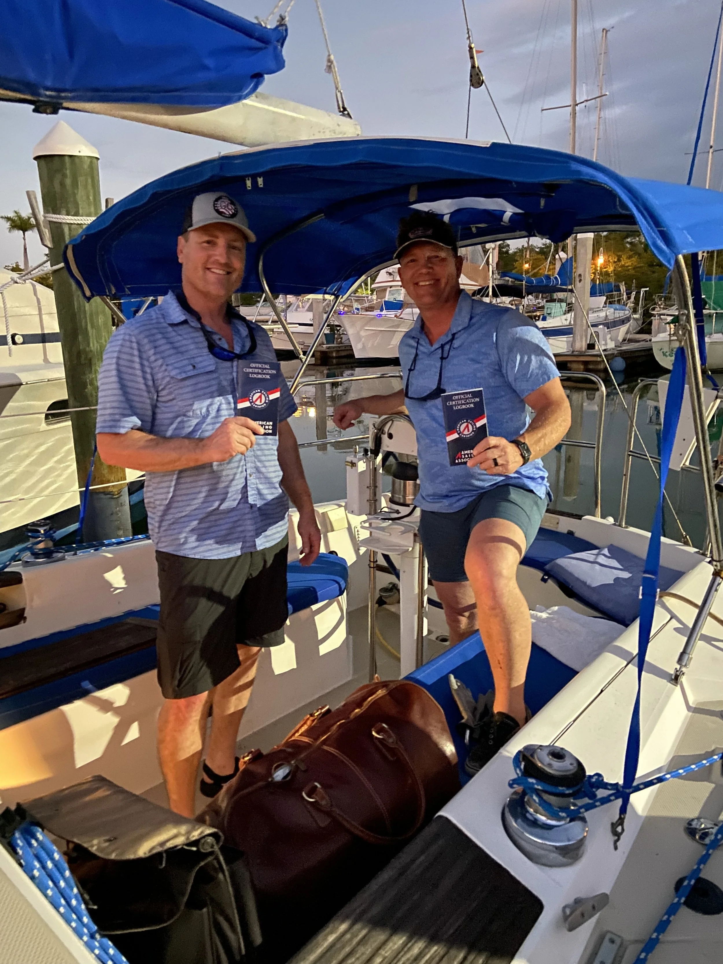 Two men on a sailboat at a marina, holding official certification books, smiling, with sailboats docked in the background during evening time.