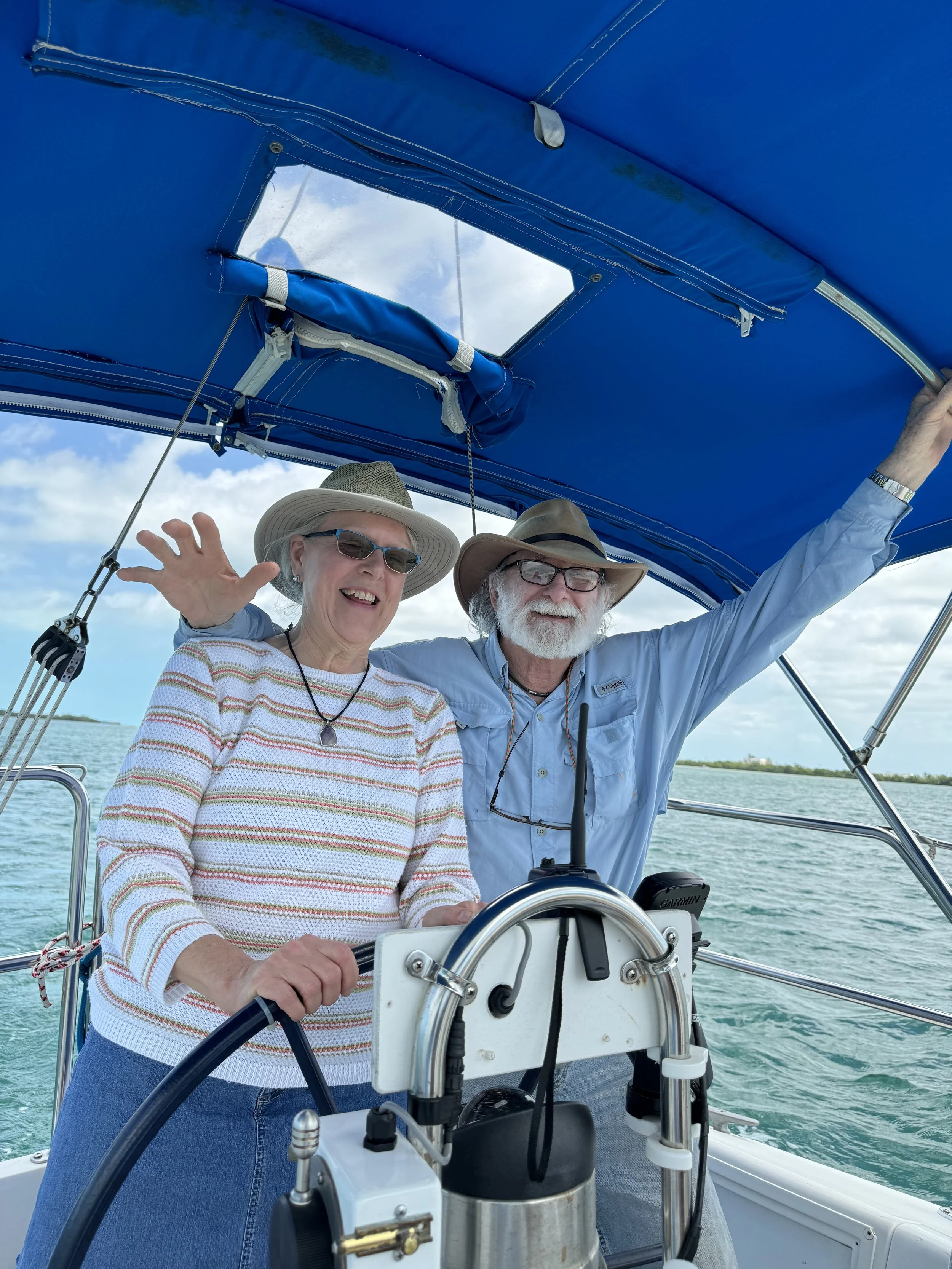 An elderly couple sailing on a boat, smiling, wearing hats and sunglasses, with a view of the water and sky in the background.