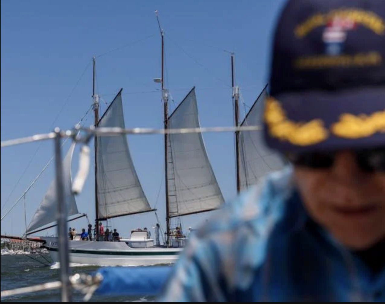 A veteran man in a navy hat and plaid shirt on a boat, with a two-masted sailboat and its sails in the background on a clear day.