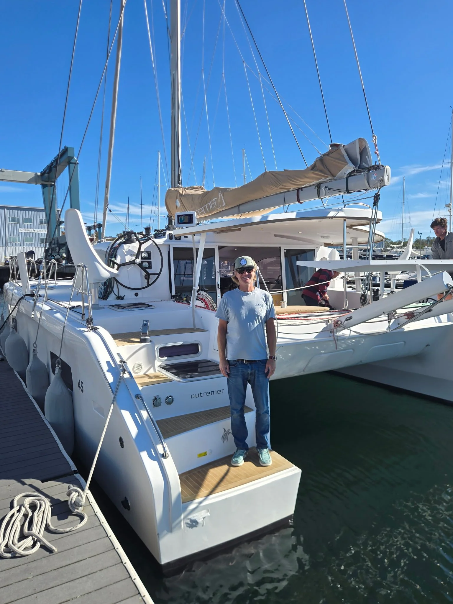 A woman standing on the back deck of a white sailboat named Outremer, docked at a marina with other boats and yachts around, under a clear blue sky.