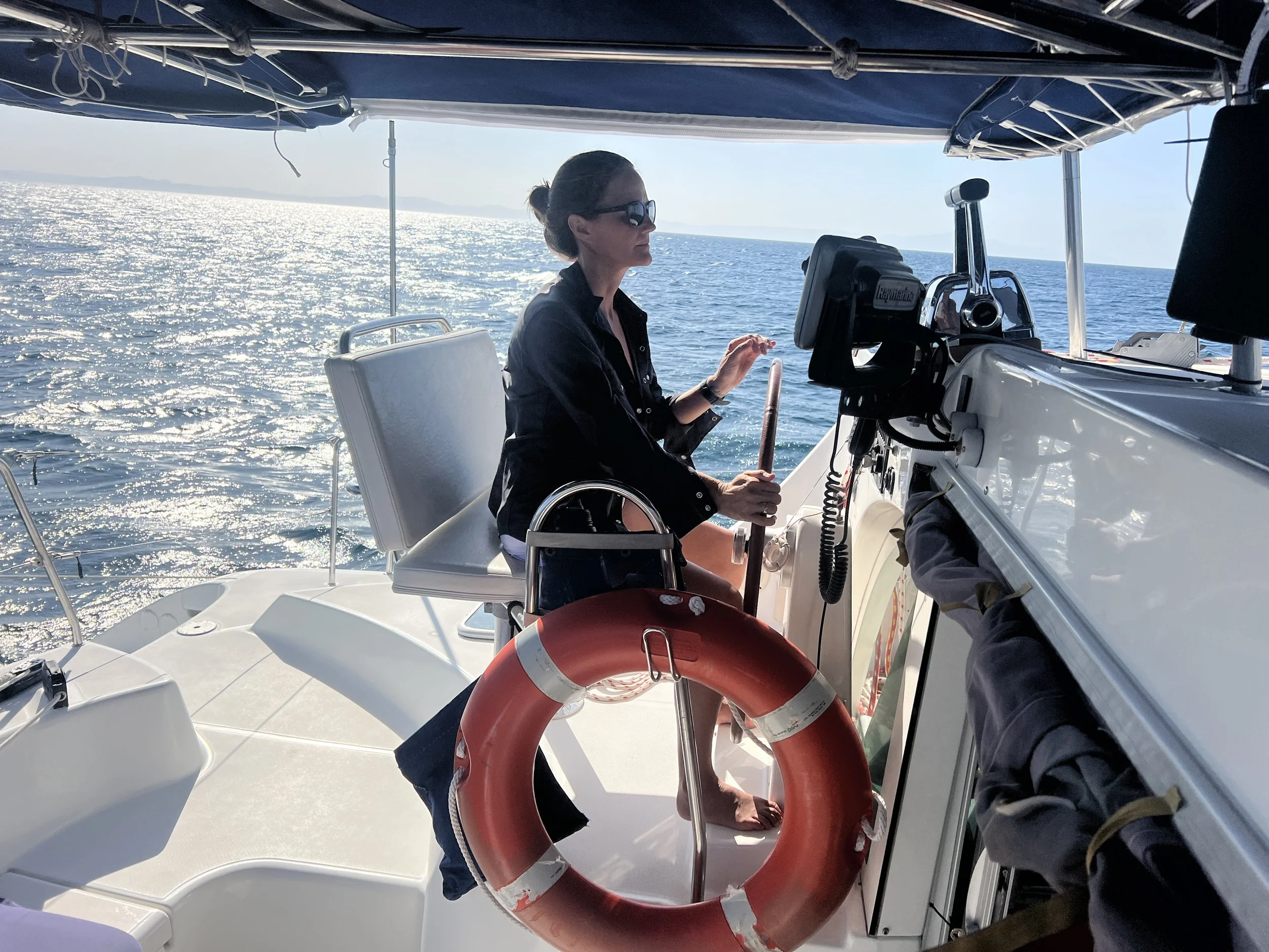A woman in sunglasses and a black jacket steering a boat on the water with sun reflecting off the ocean, wearing a life preserver hanging on the boat.