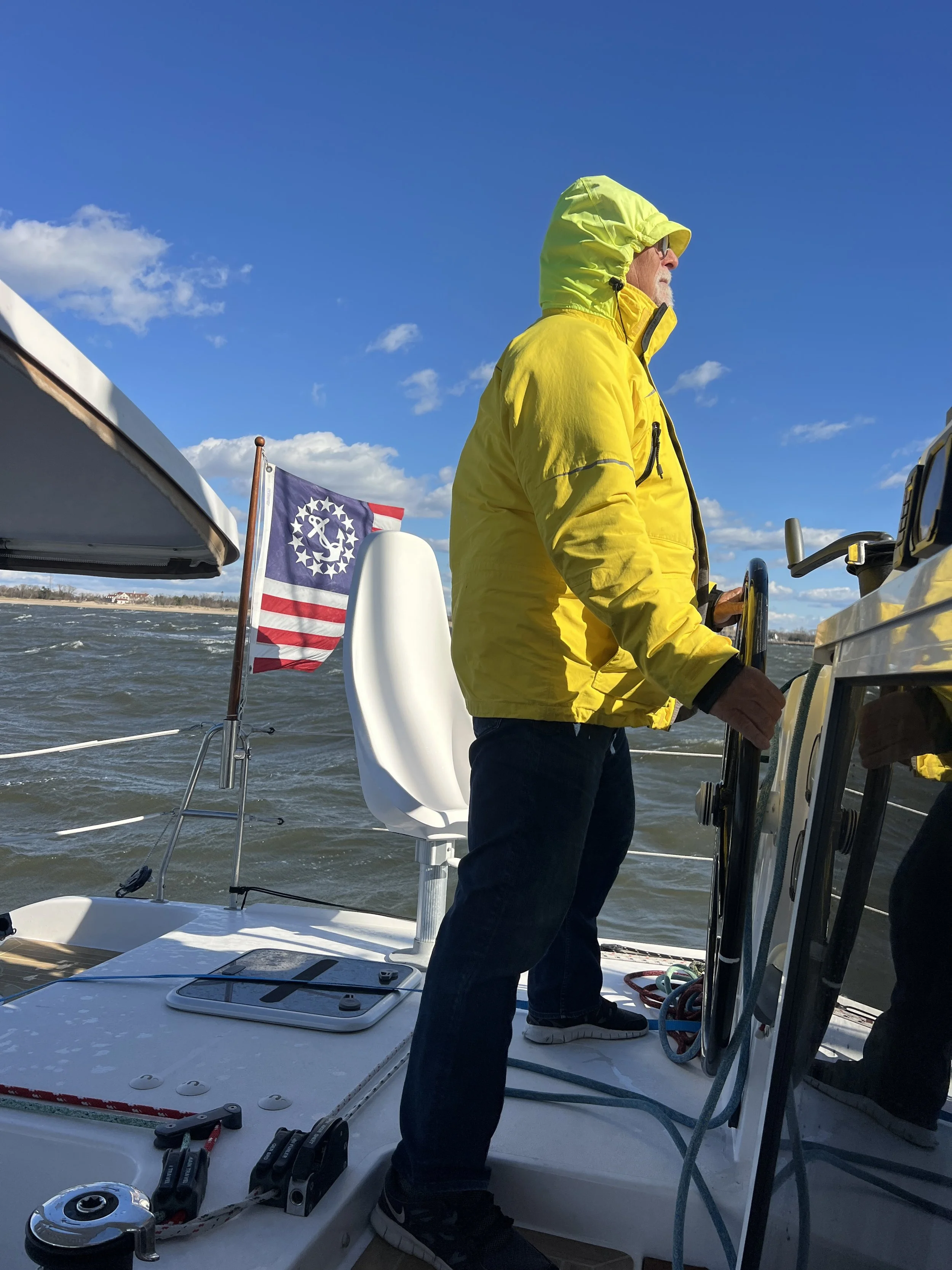 Man in yellow jacket steering a sailboat with an American flag in the background on a partly cloudy day.
