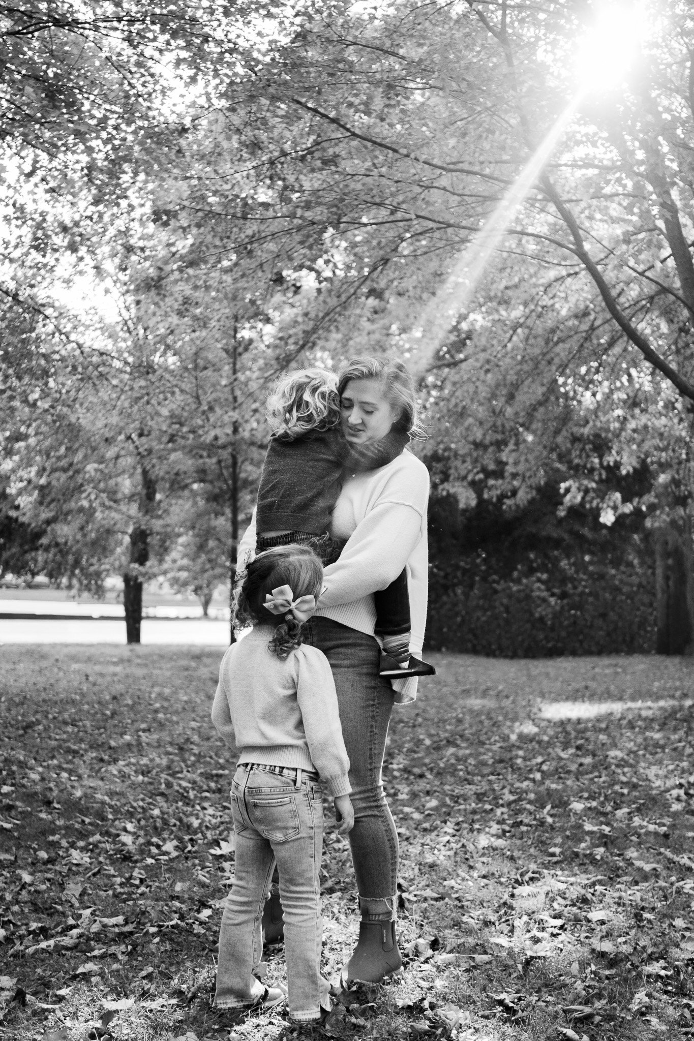 A woman hugging a small child in a park during the daytime, with another girl standing nearby watching them, surrounded by trees and fallen leaves, sunlight streaming through the branches.