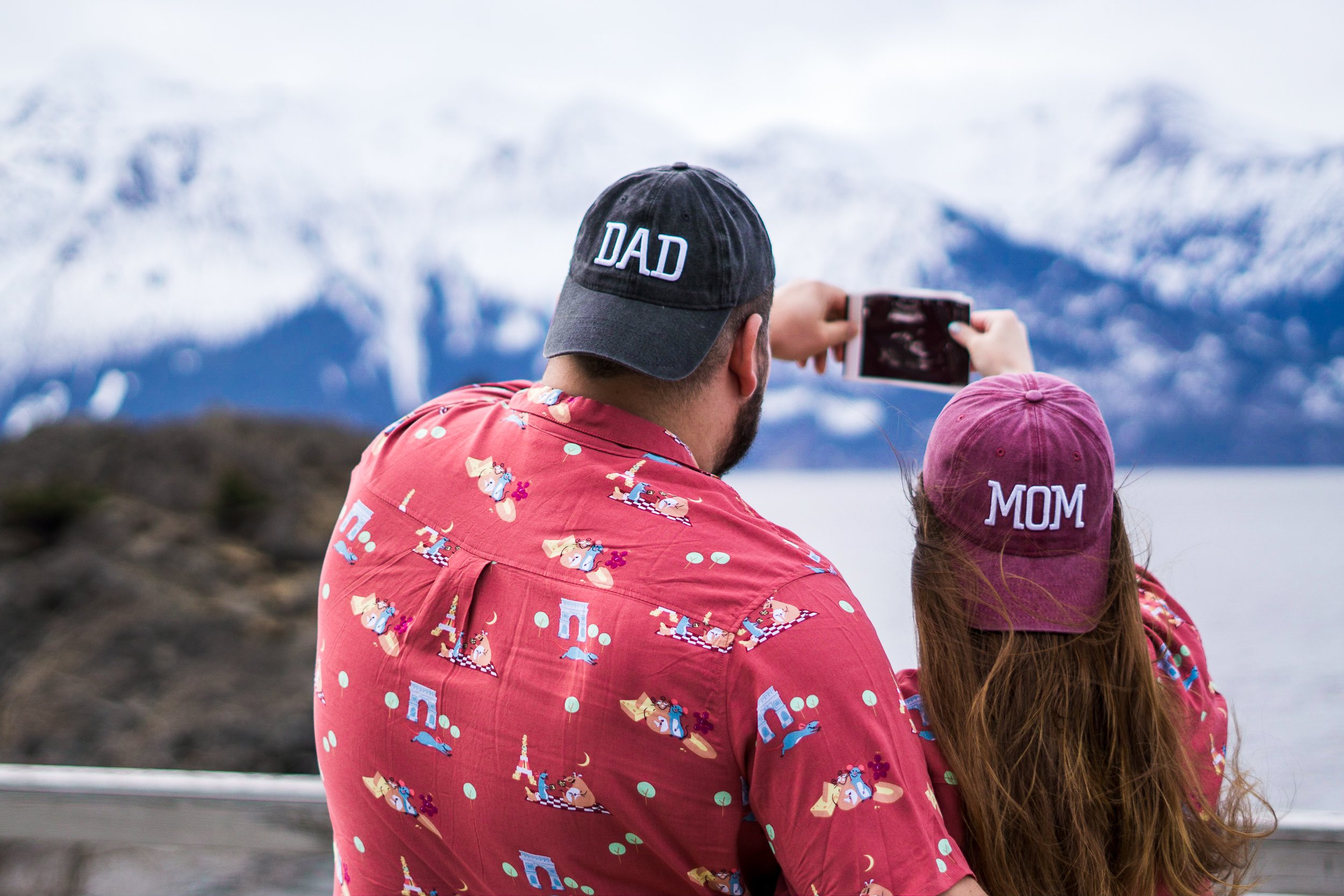 A man and a woman are taking a photo in front of snow-capped mountains and a lake. The man is wearing a dark cap with 'DAD' on it and a red shirt with playful patterns. The woman is wearing a pink cap with 'MOM' on it.