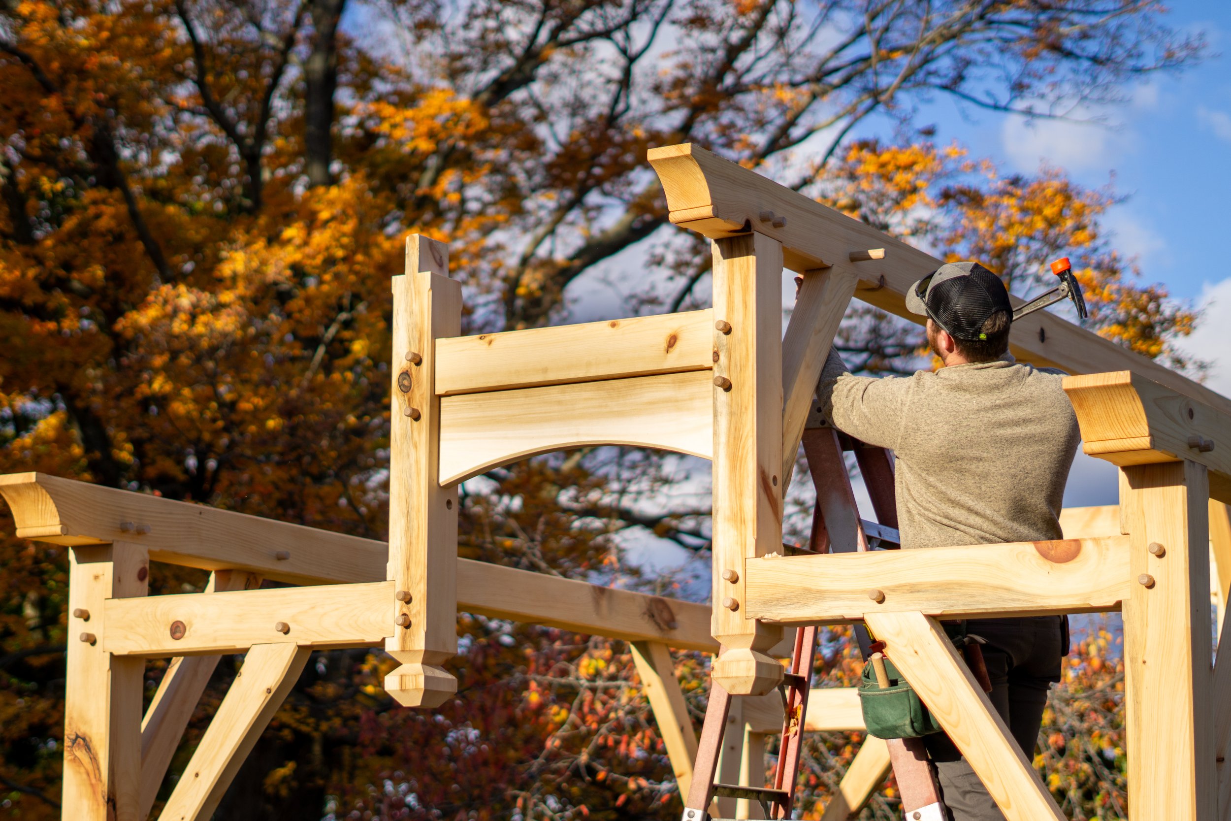 A man constructing a wooden deck outdoors during autumn, with colorful fall leaves and partly cloudy sky in the background.