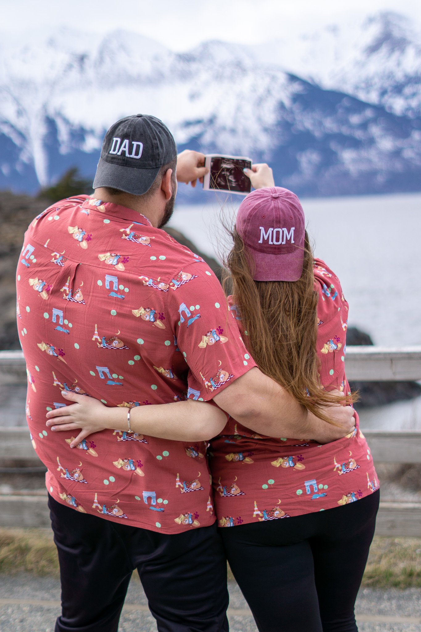 A man and woman, both wearing matching pink shirts and hats labeled 'Dad' and 'Mom', are taking a photo of snow-capped mountains and a body of water in the background.