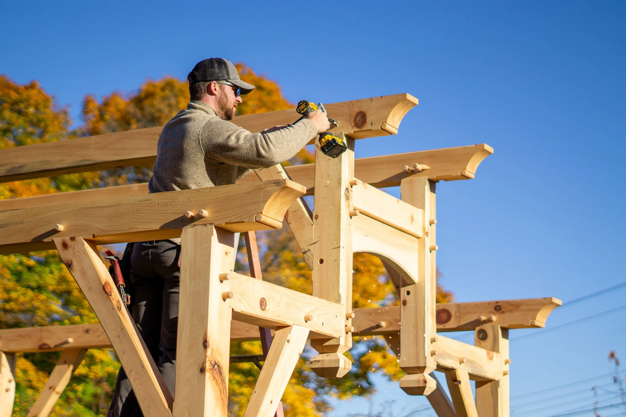 A man constructing a wooden structure outdoors with a power drill, surrounded by trees with autumn leaves, under a clear blue sky.