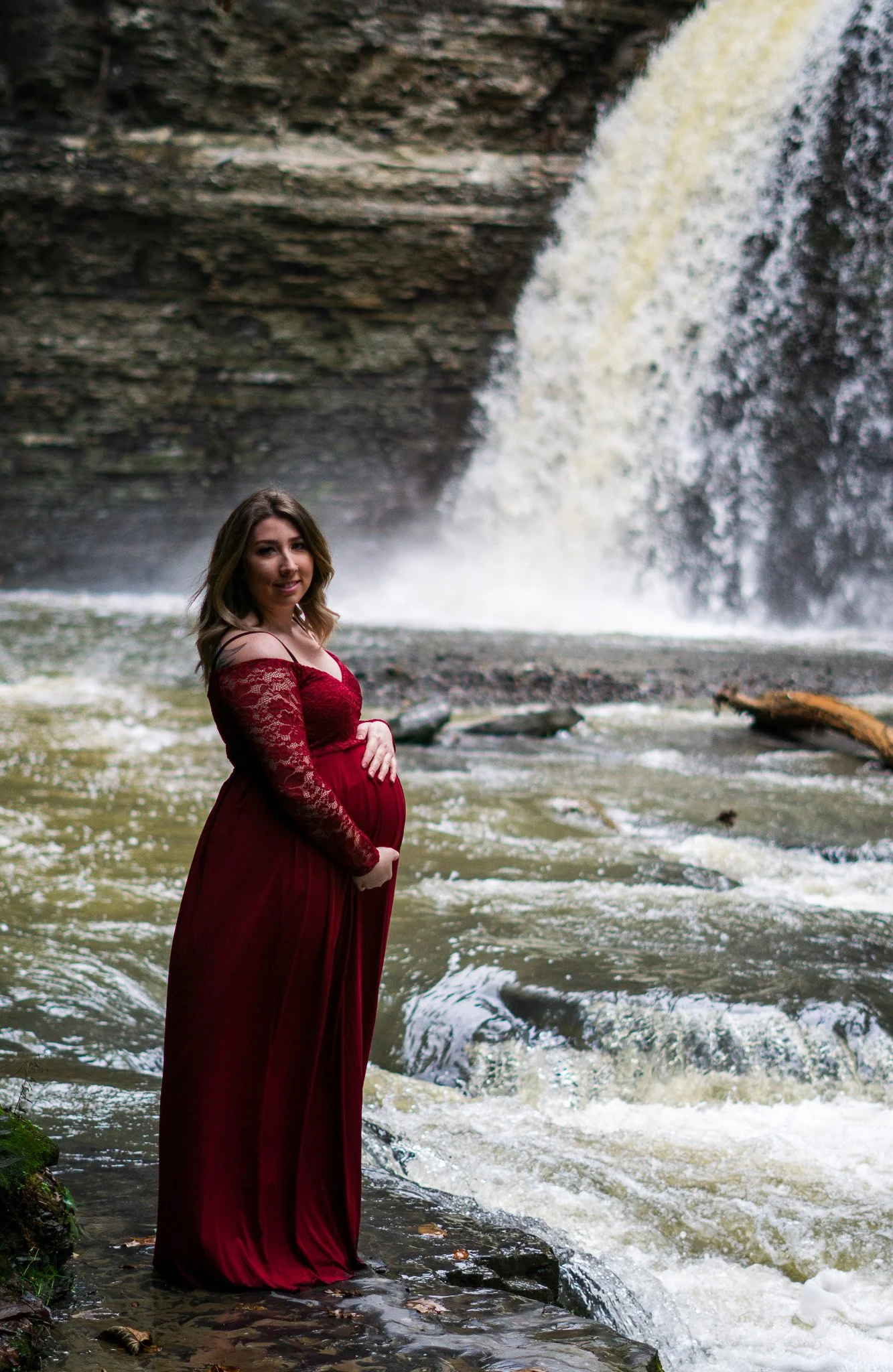 Pregnant woman in a red dress standing on rocks near a waterfall.