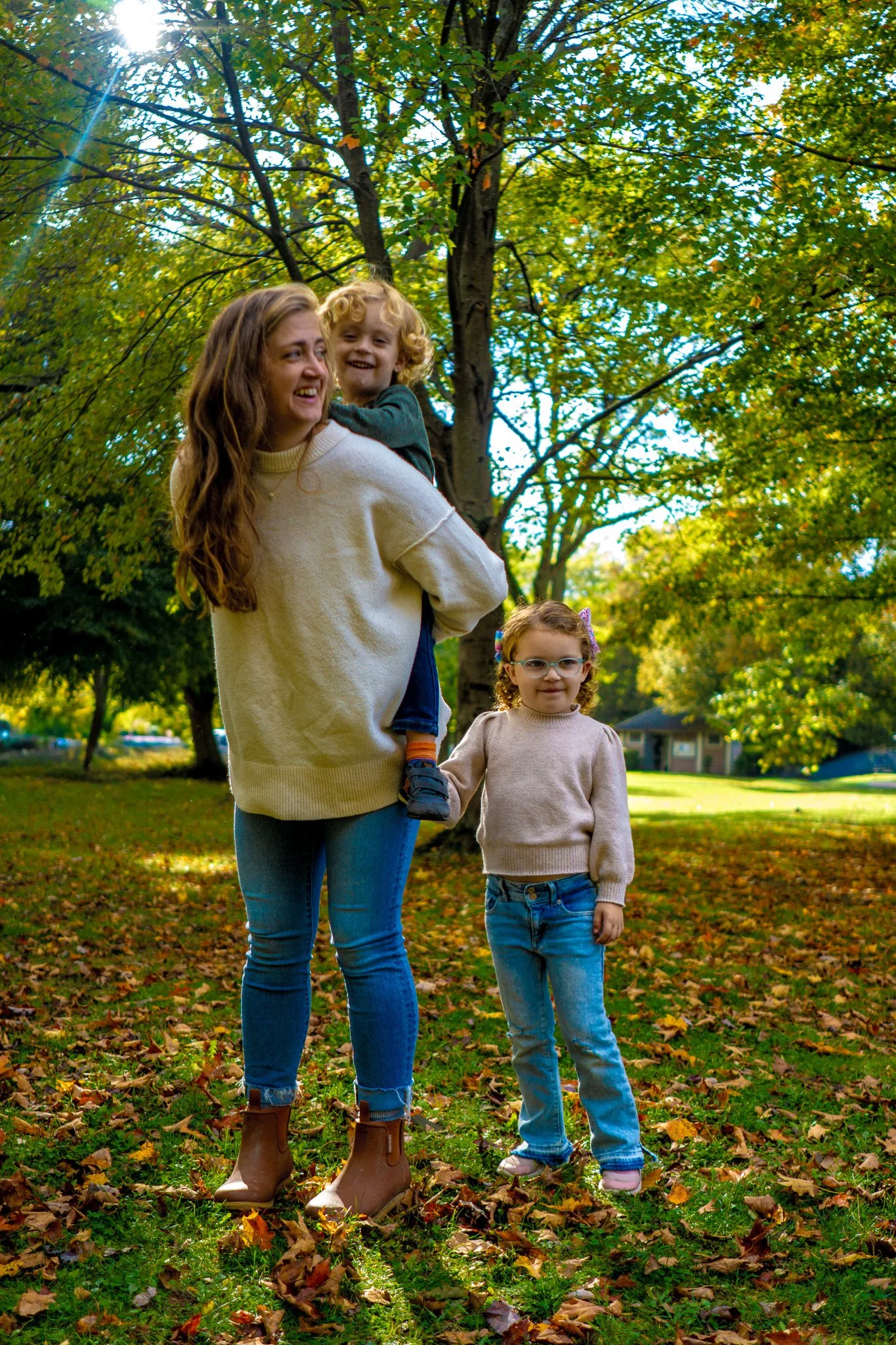 A woman with long brown hair is holding a young boy with curly blond hair on her shoulder, both smiling. A young girl with glasses and curly hair stands beside them holding her mother's hand, in a park with trees and fallen autumn leaves.