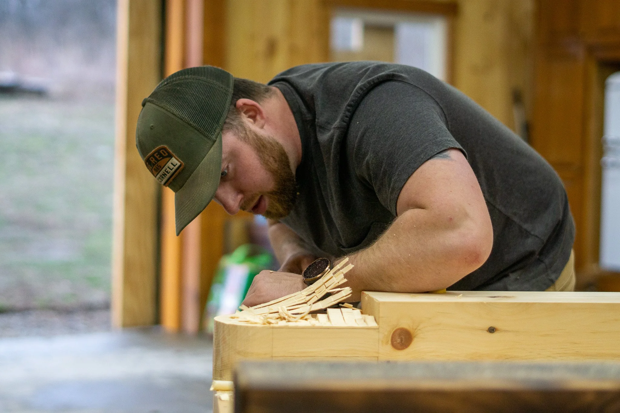 A man with a beard and wearing a cap is working on a piece of wood. He is marking or carving it while leaning over a workbench in a woodworking shop.