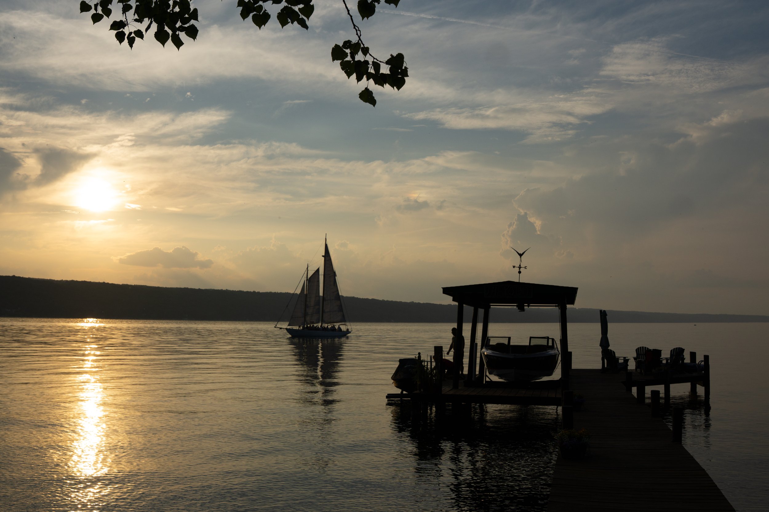 Silhouette of a woman on a dock with a boat, a sailboat on the water, and a sunset over the lake.