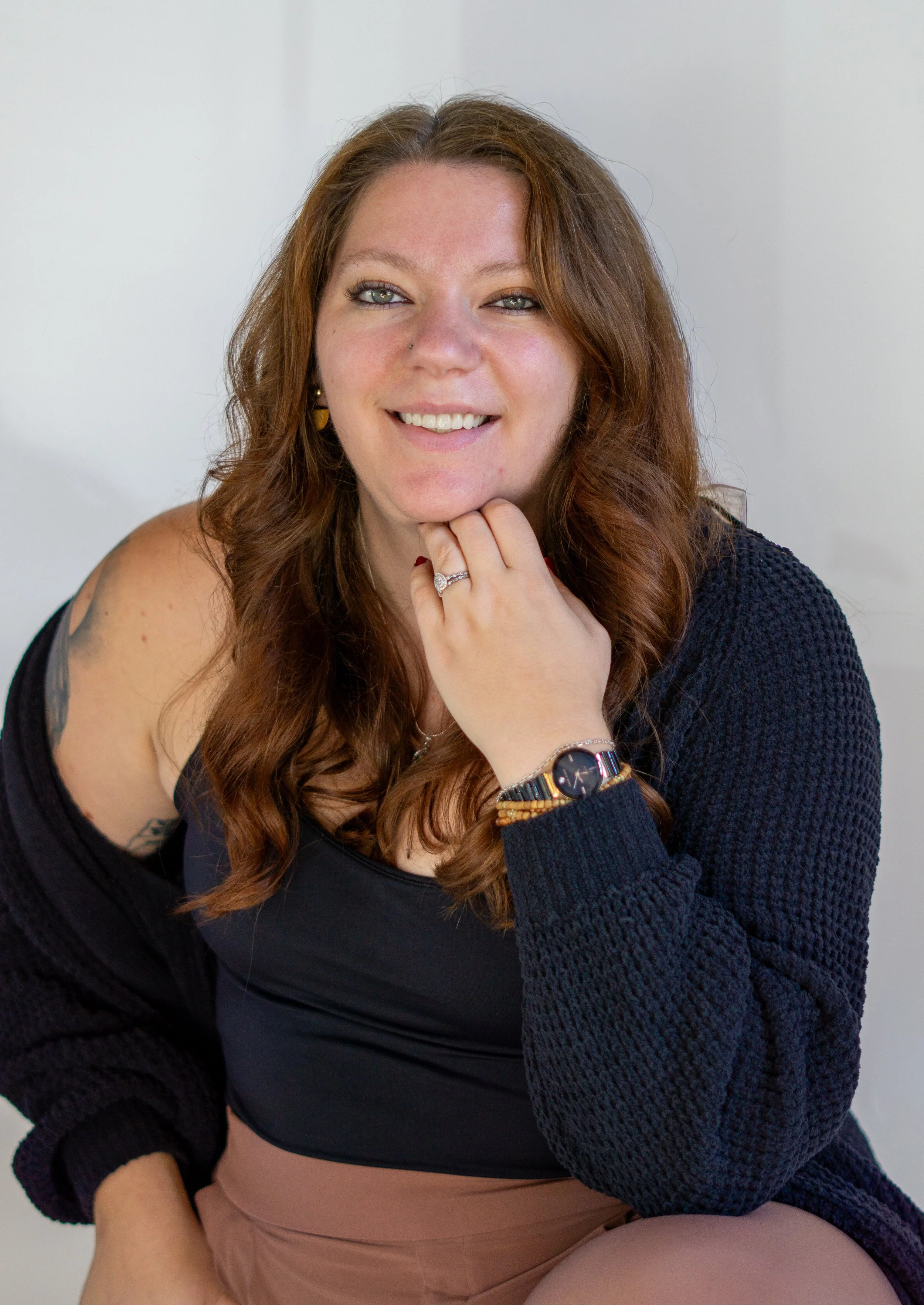 A woman with red hair, blue eyes, and a cheerful smile, wearing a black tank top, beige pants, a black cardigan, a watch, and jewelry, sitting against a plain light-colored background.