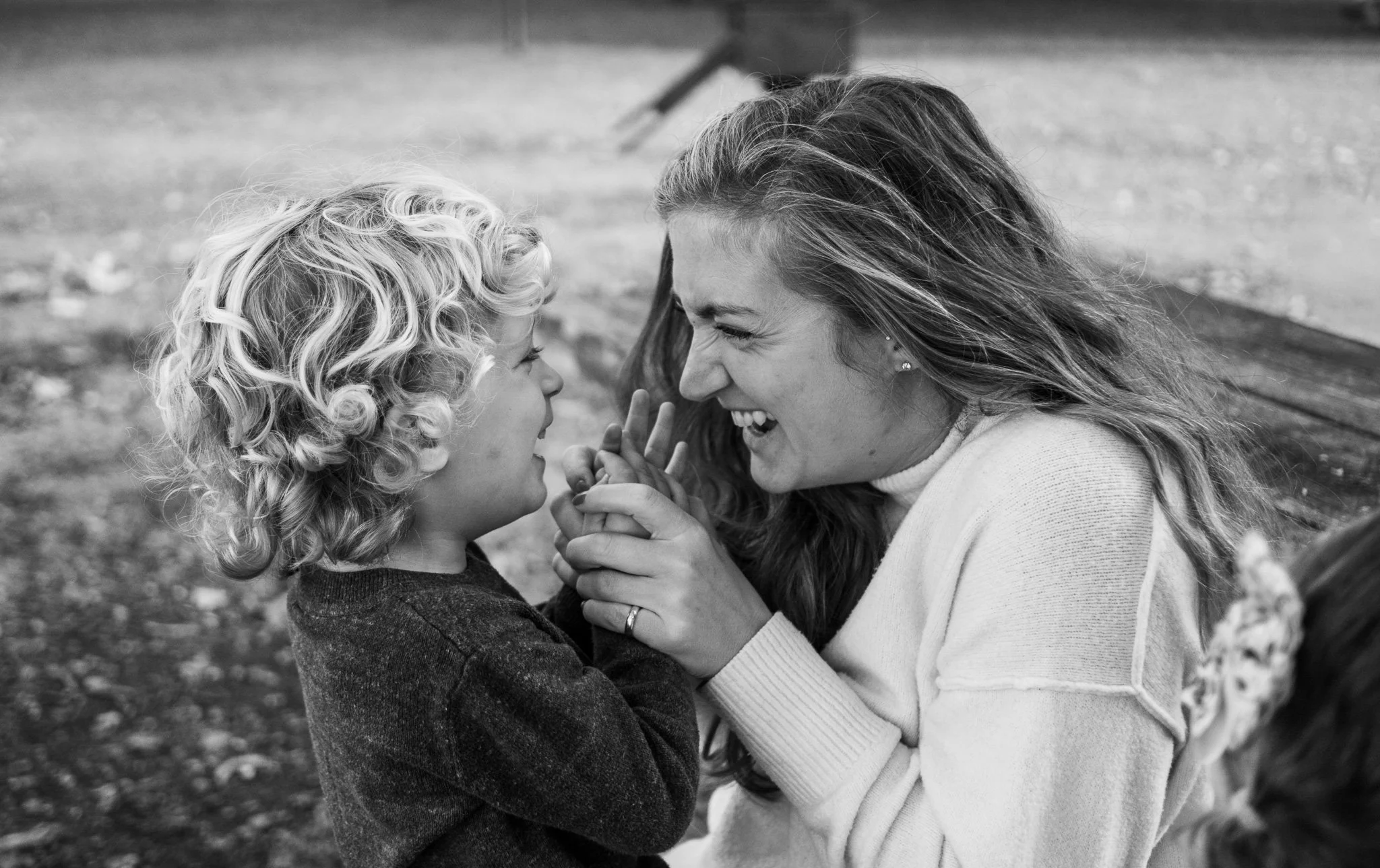 A woman and a young boy face each other closely, smiling and holding hands, outdoors with a blurred background.