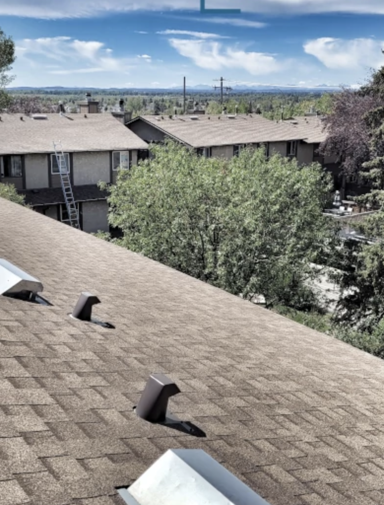 View from a rooftop showing neighboring apartment buildings, trees, and a distant landscape with mountains under a partly cloudy sky.