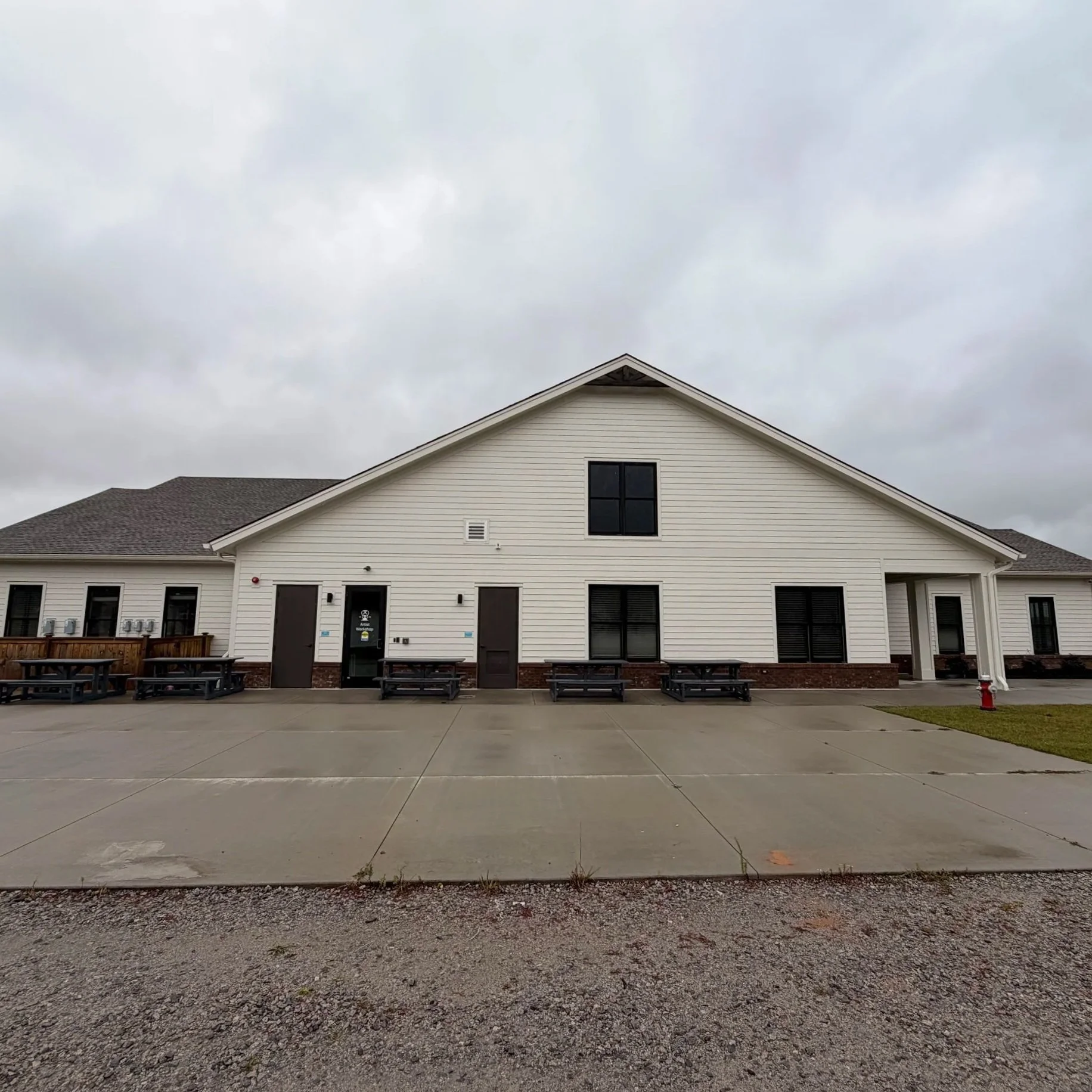 Front view of a modern, white building with a gable roof, multiple windows, and a small covered entryway. There are benches and a fire hydrant near the sidewalk under overcast skies.