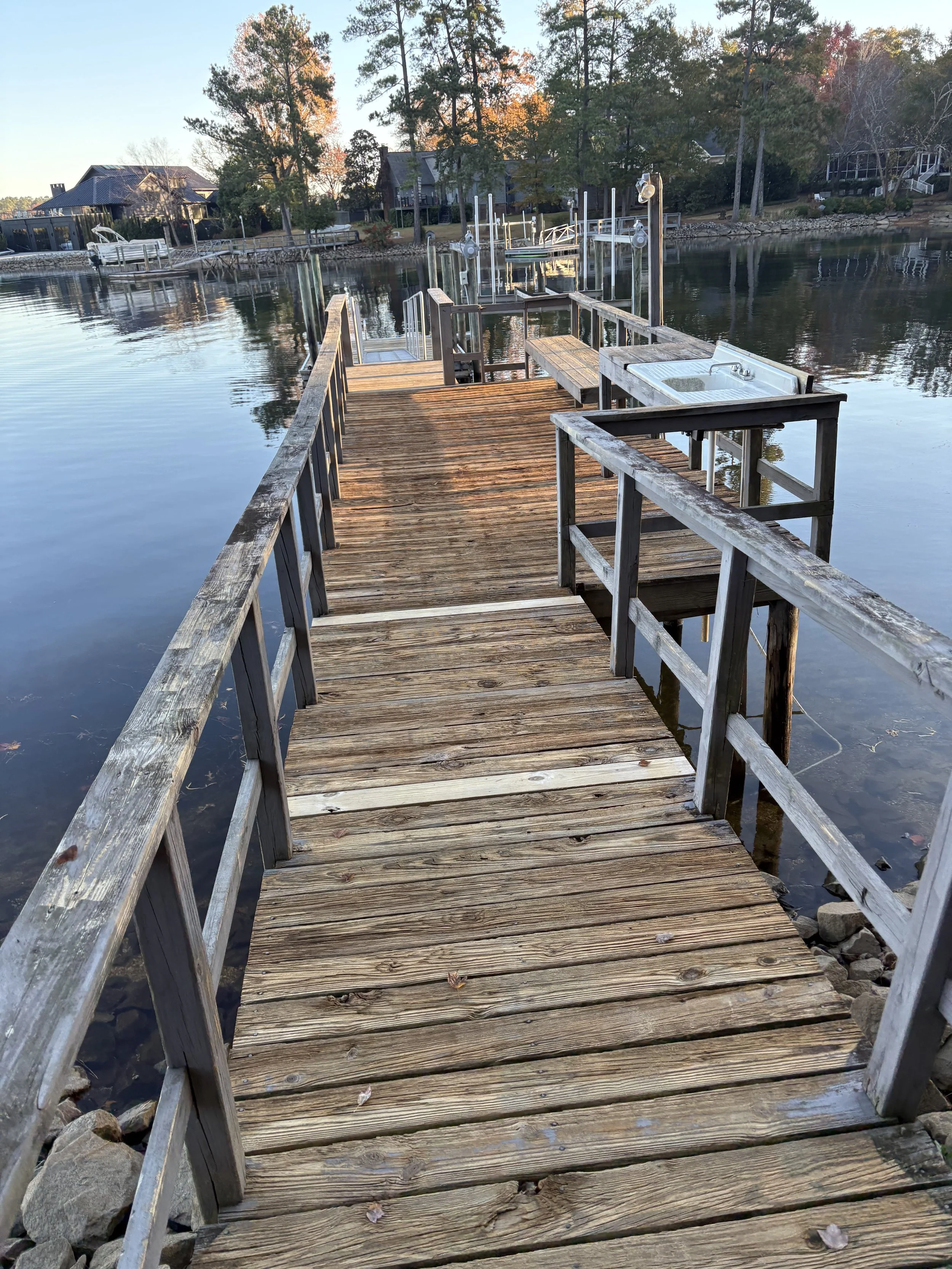 A wooden dock extending over calm water with trees and houses in the background. The dock has railings, a sink, and several boat slips.