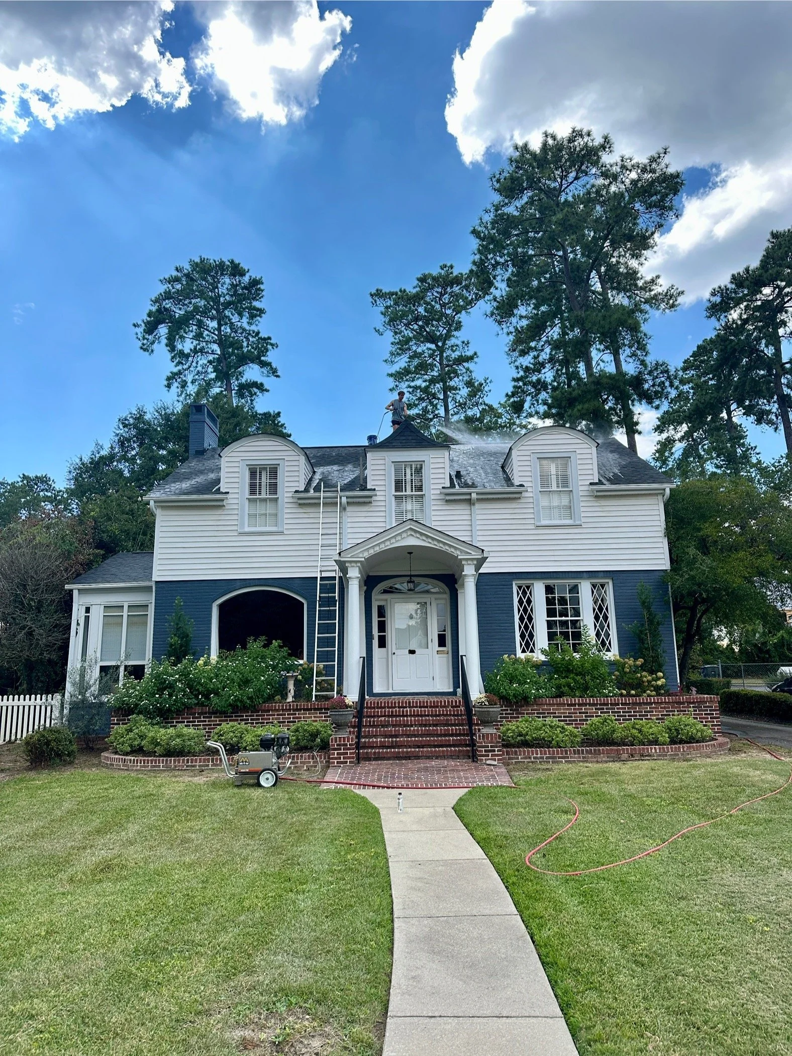 A large two-story house with blue and white exterior, brick stairs leading to the front door, and surrounded by a well-maintained lawn and bushes. A person is cleaning the roof on a ladder, and a hose is watering the roof.