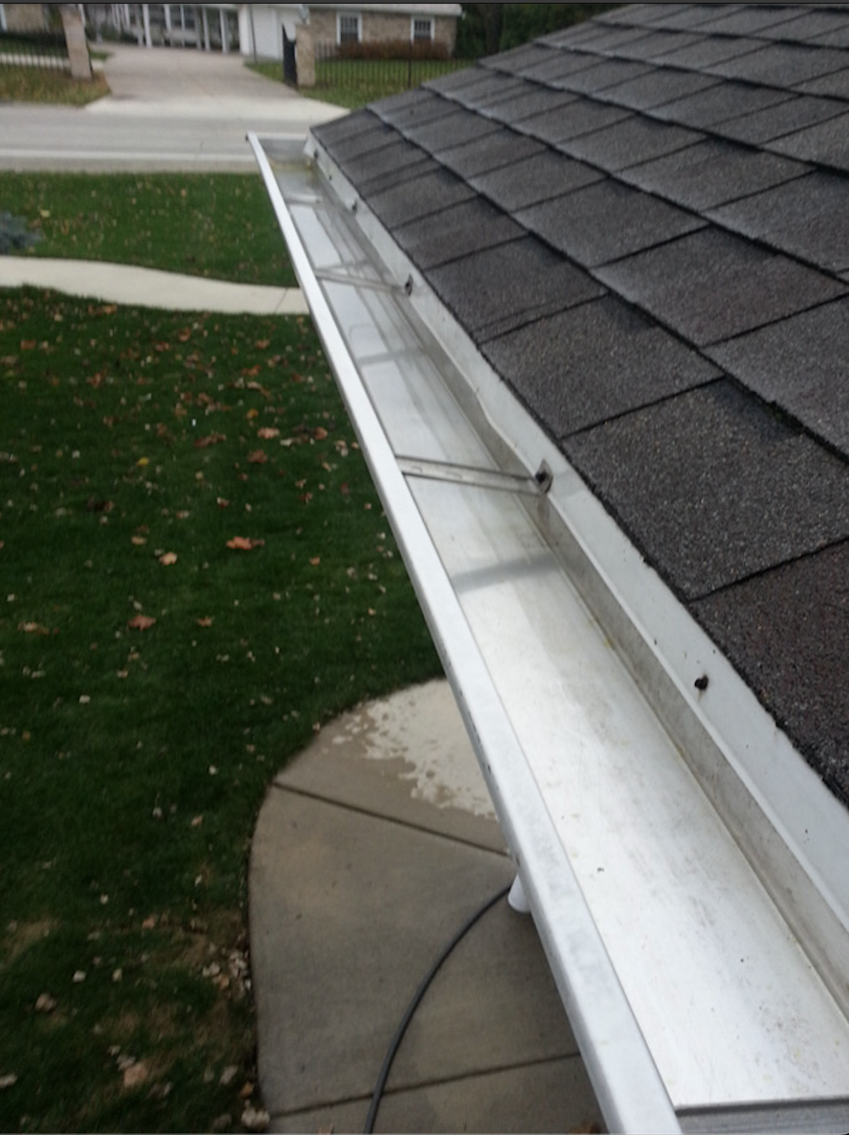 View of a roof's gutter with a downspout, with asphalt shingles on the roof and a yard with green grass, a sidewalk, and a street in the background.
