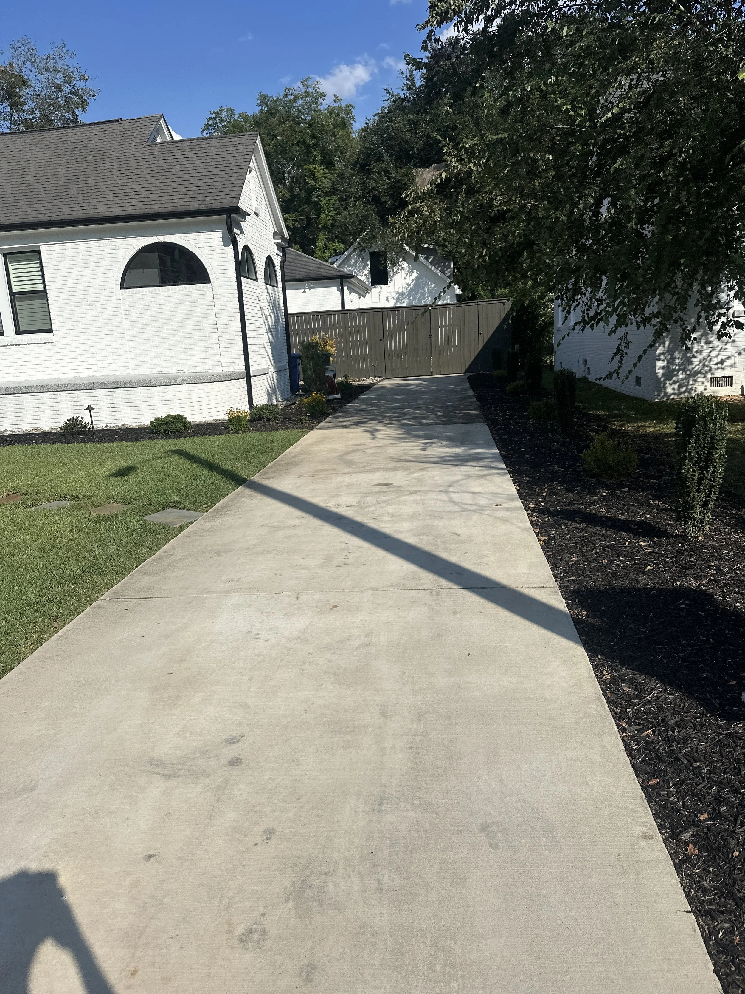 Concrete sidewalk leading to a backyard, with a white house on the left and a fence at the end of the path. There are trees and a garden bed with small plants on the right side, and a green lawn on the left side. The sky is blue with some clouds.