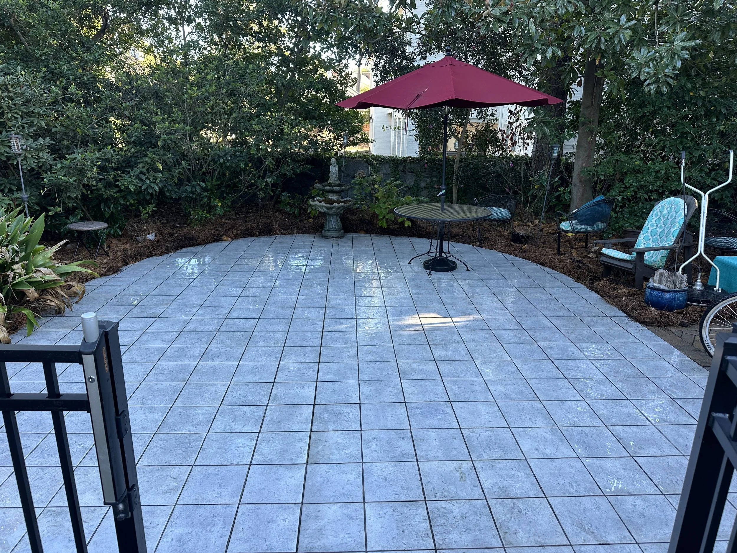 Back patio with light blue tile flooring, a red umbrella over a round table, and three chairs, surrounded by dense green bushes and trees.