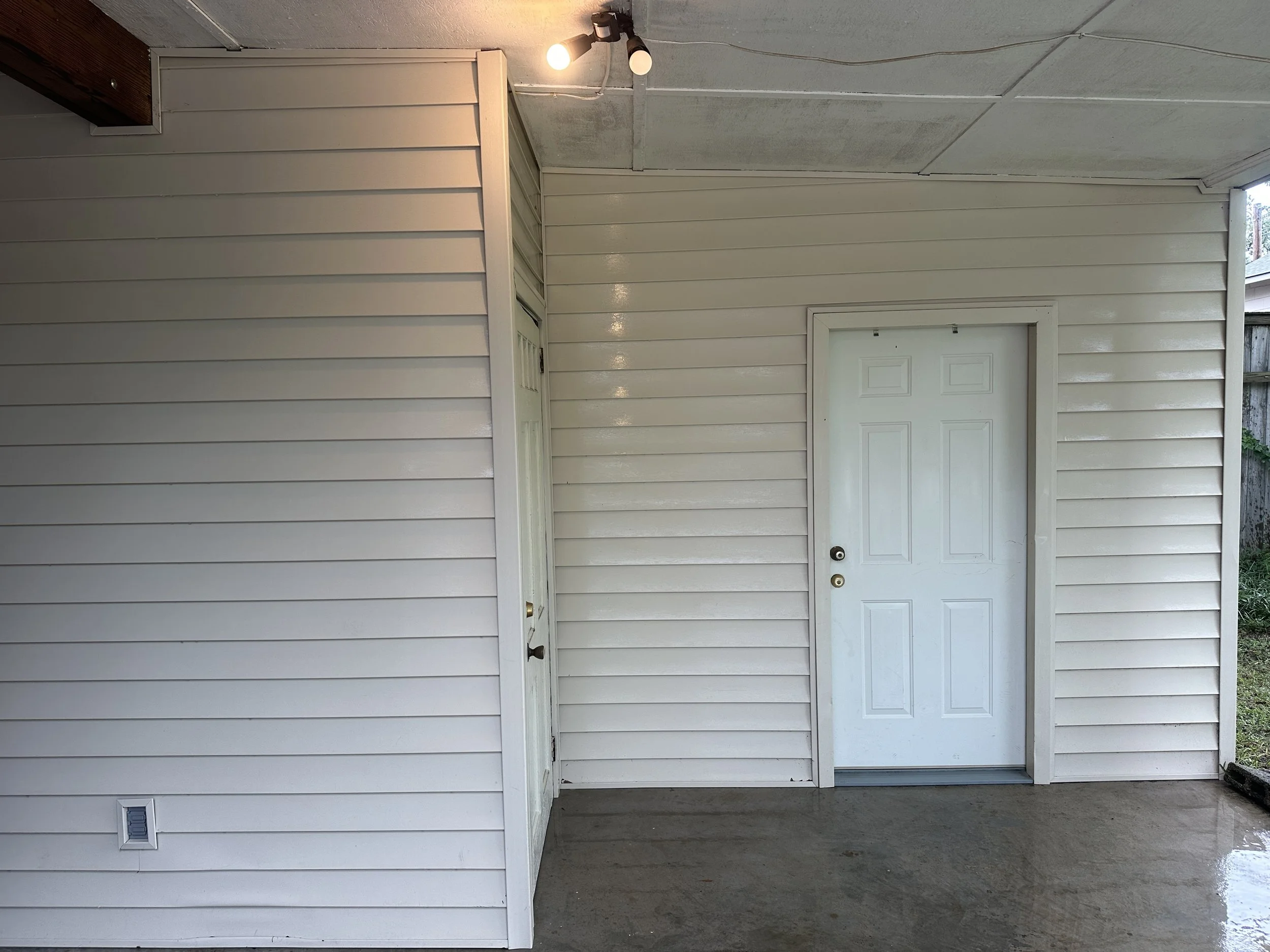 View of a covered porch with white vinyl siding walls, a white door with a deadbolt lock, an overhead light fixture, and a small vent near the floor.