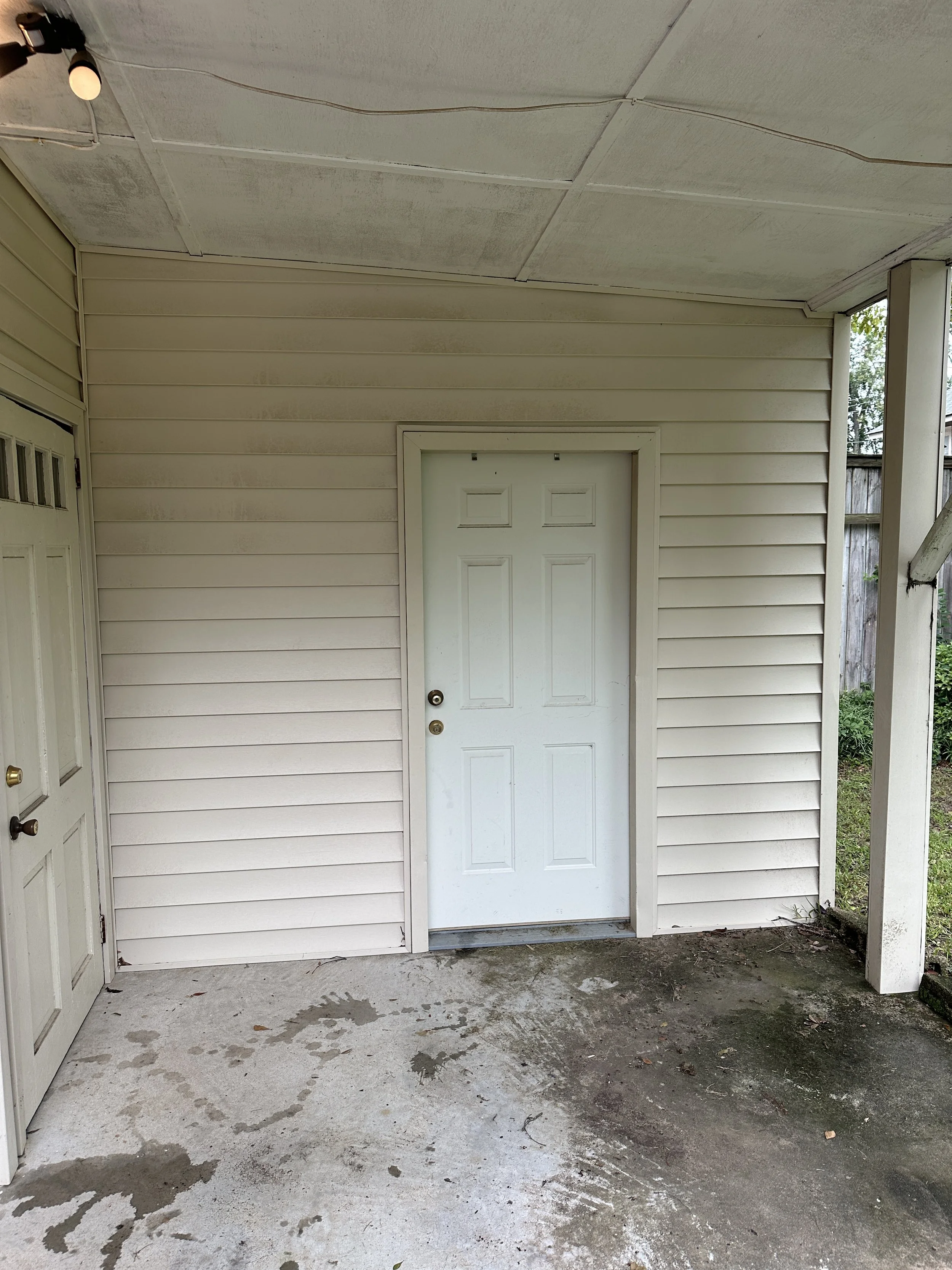 Outside porch area with an off-white vinyl siding wall, a white door in the center, and a partially visible door to the left. The concrete floor has wet patches and dirt.