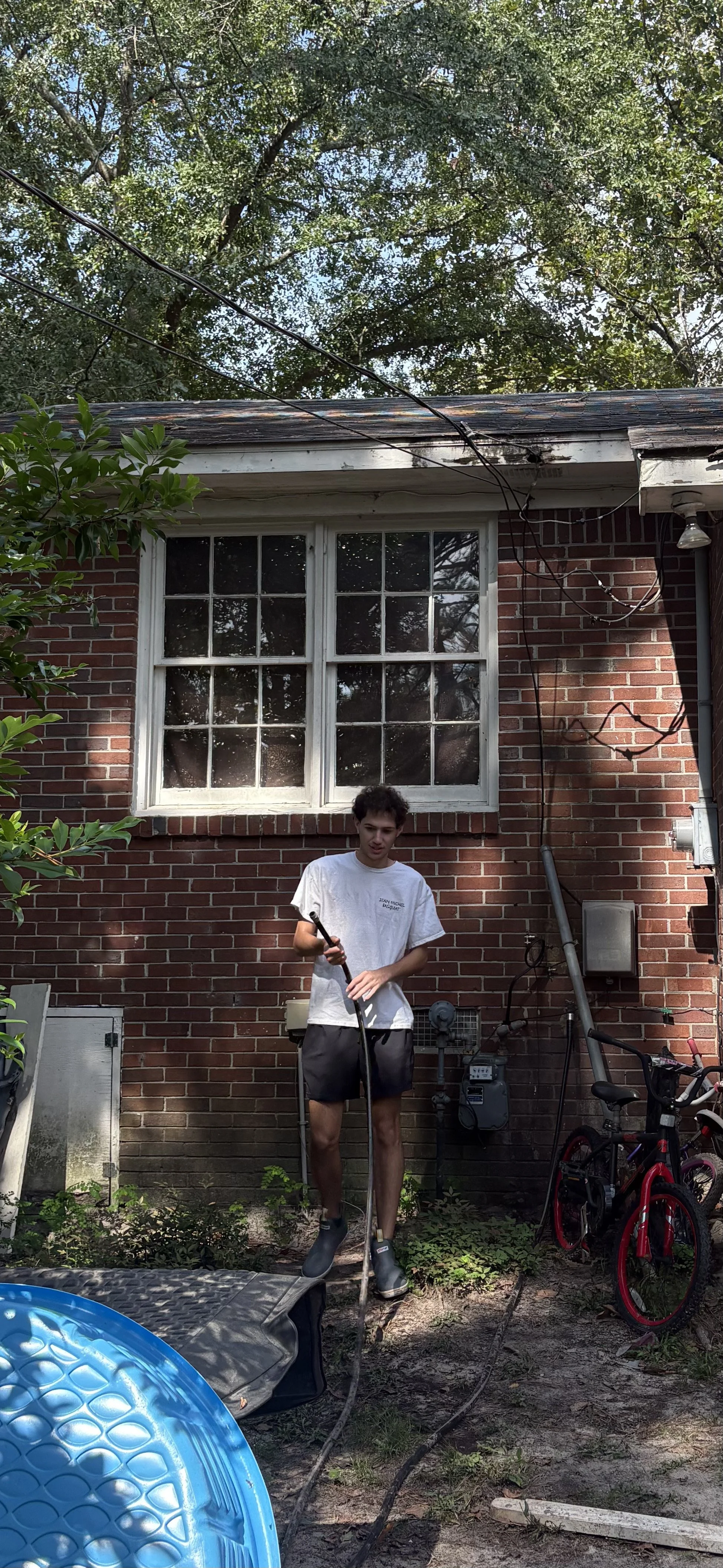 A young man standing outdoors in a backyard, holding a garden hose, wearing a white t-shirt, black shorts, and slip-on shoes, with bicycles, a small pool, and a brick house with a window in the background.