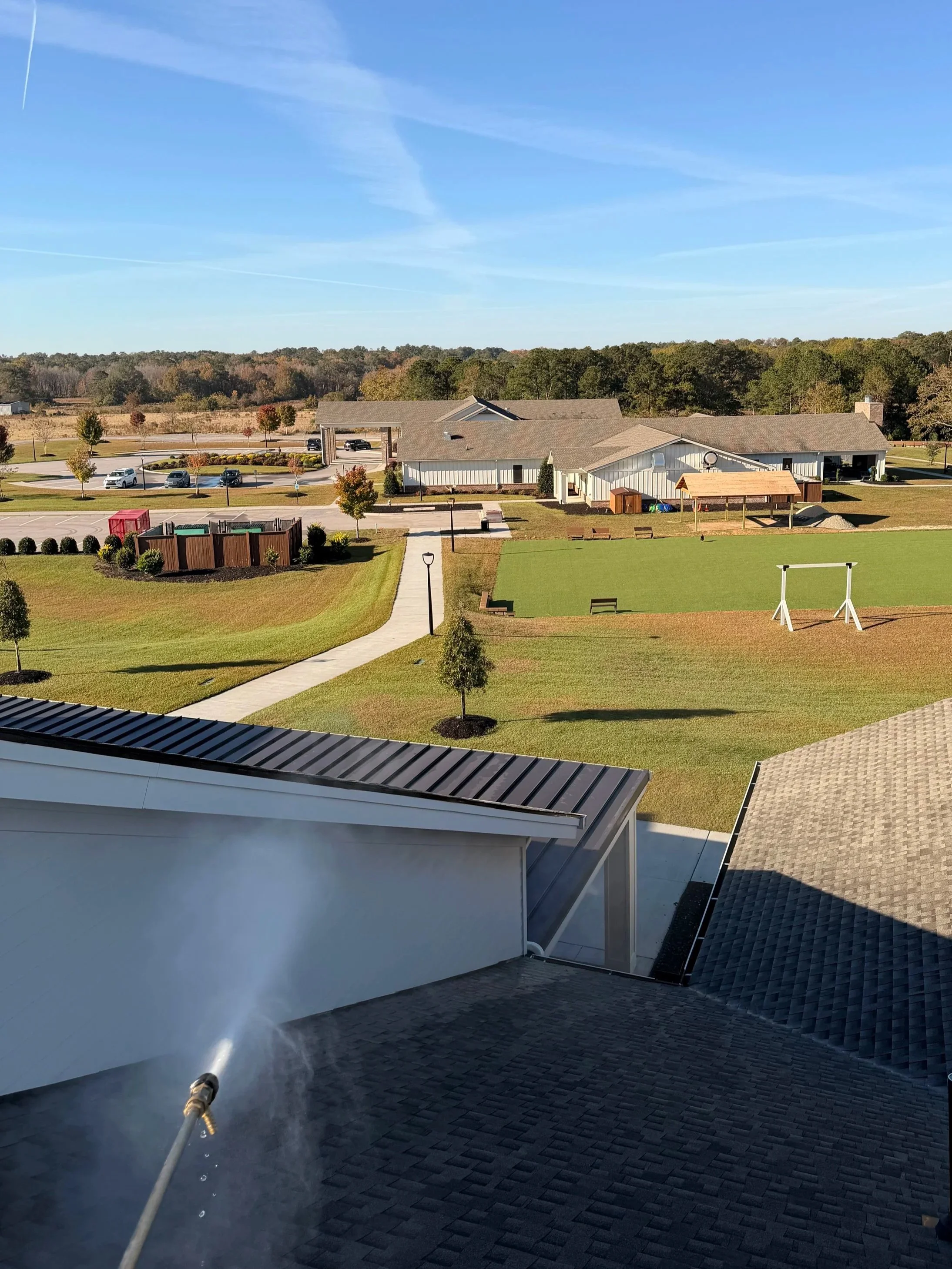 View of a school or community center campus with green lawns, pathways, trees, parking lot with cars, and buildings under a clear blue sky.