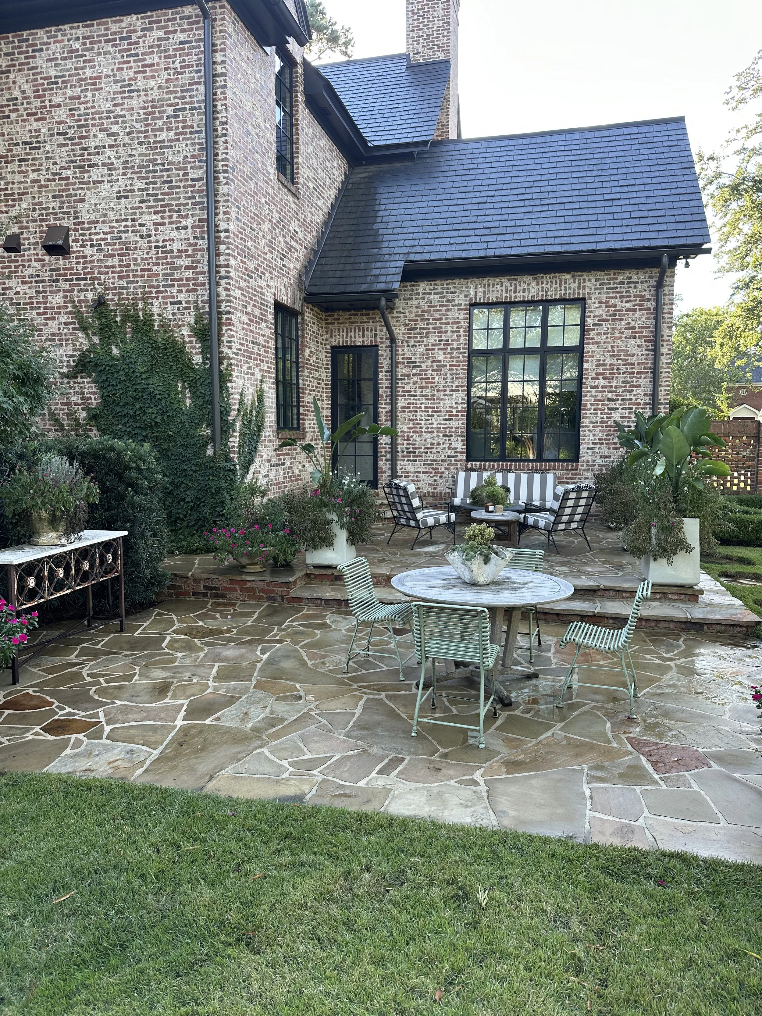 A brick house with a patio area, featuring a stone floor, potted plants, and outdoor furniture including a round table with four chairs and a seating area with striped cushions on black metal chairs.