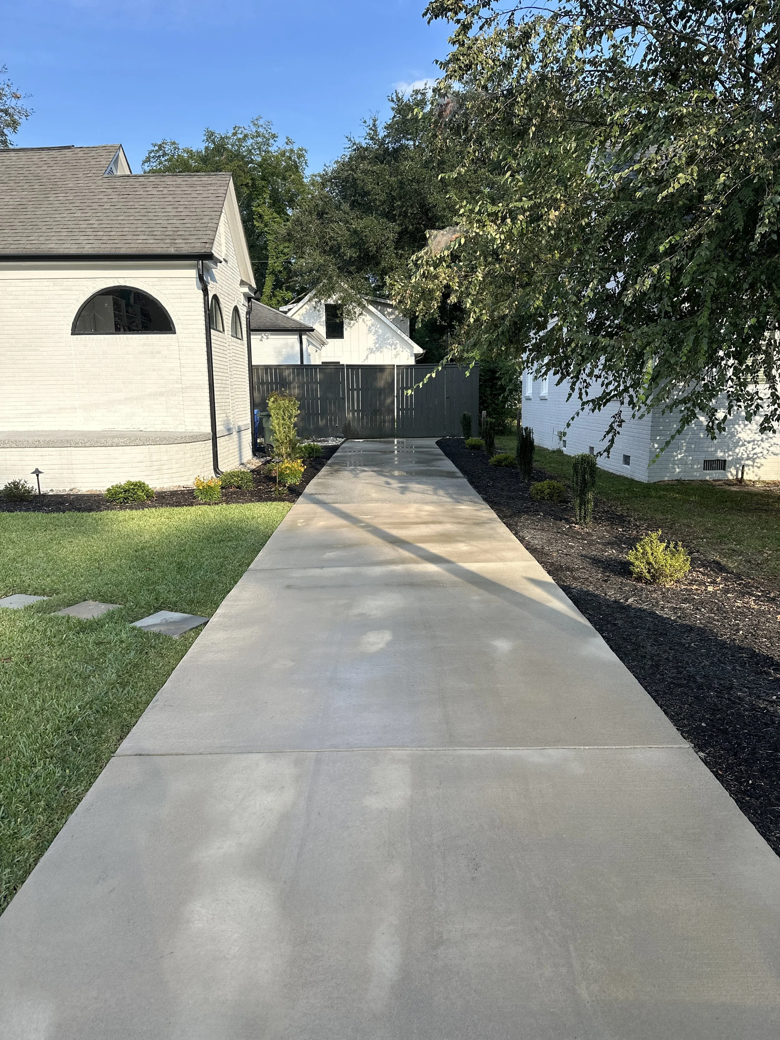 Concrete driveway leading to a black fence gate, with landscaped yard and trees on both sides, and neighboring houses visible in the background under a clear blue sky.