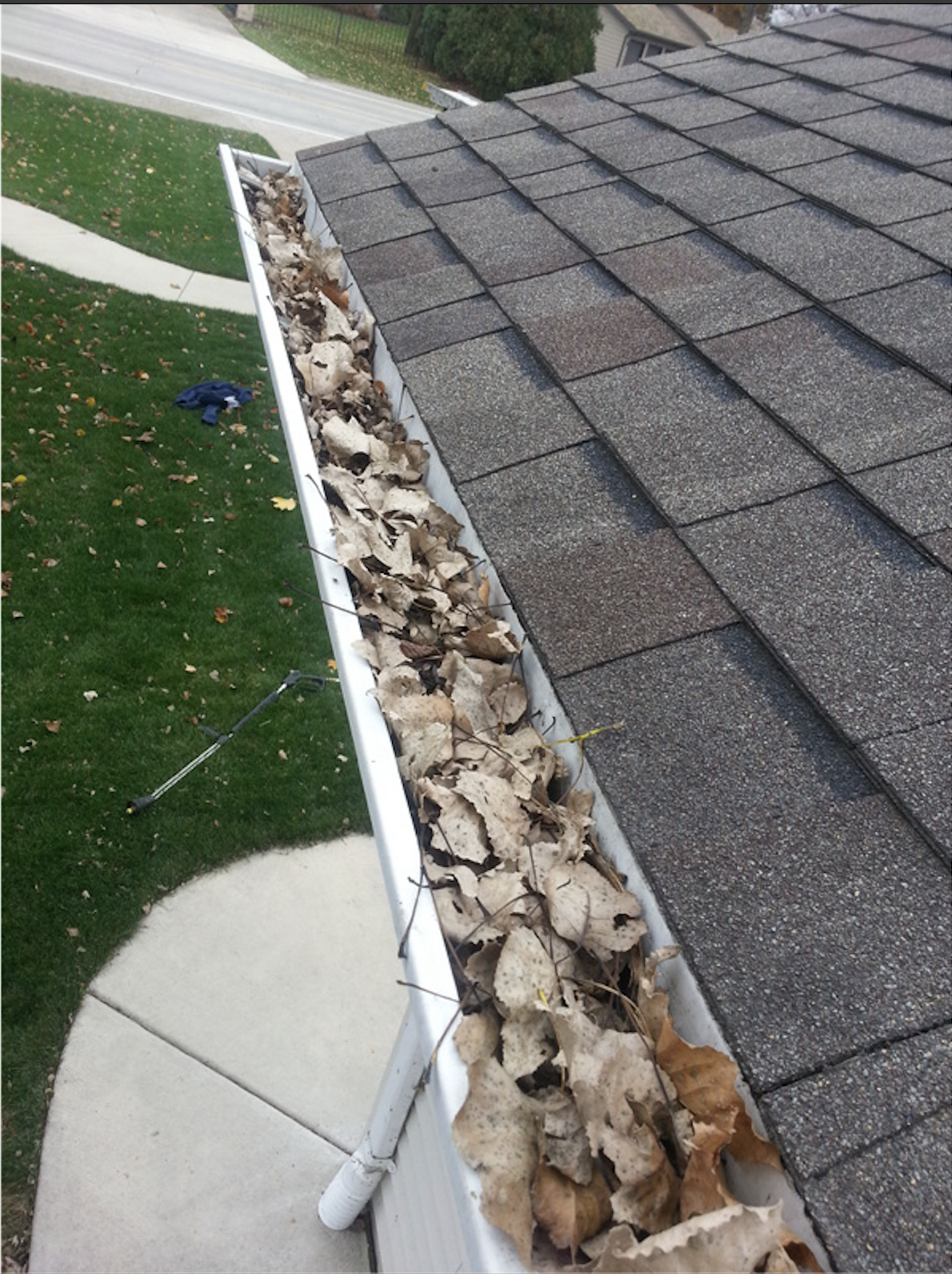 The gutter of a house filled with dry fallen leaves and debris, with shingles on the roof visible above, neighboring sidewalk, grass, and street in the background.