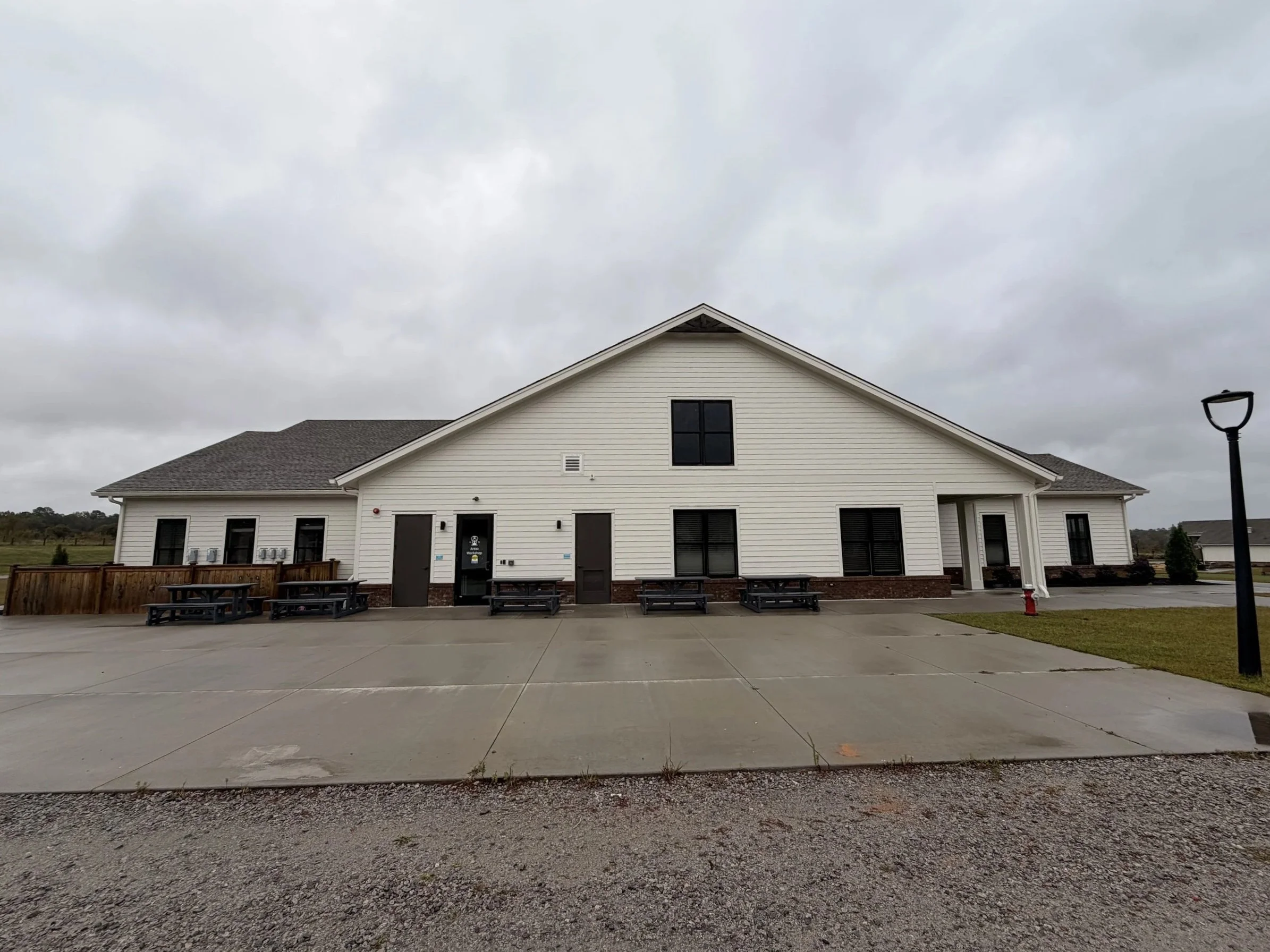 Exterior view of a white, single-story building with a gable roof, multiple windows, and several picnic tables in front, on a cloudy day.
