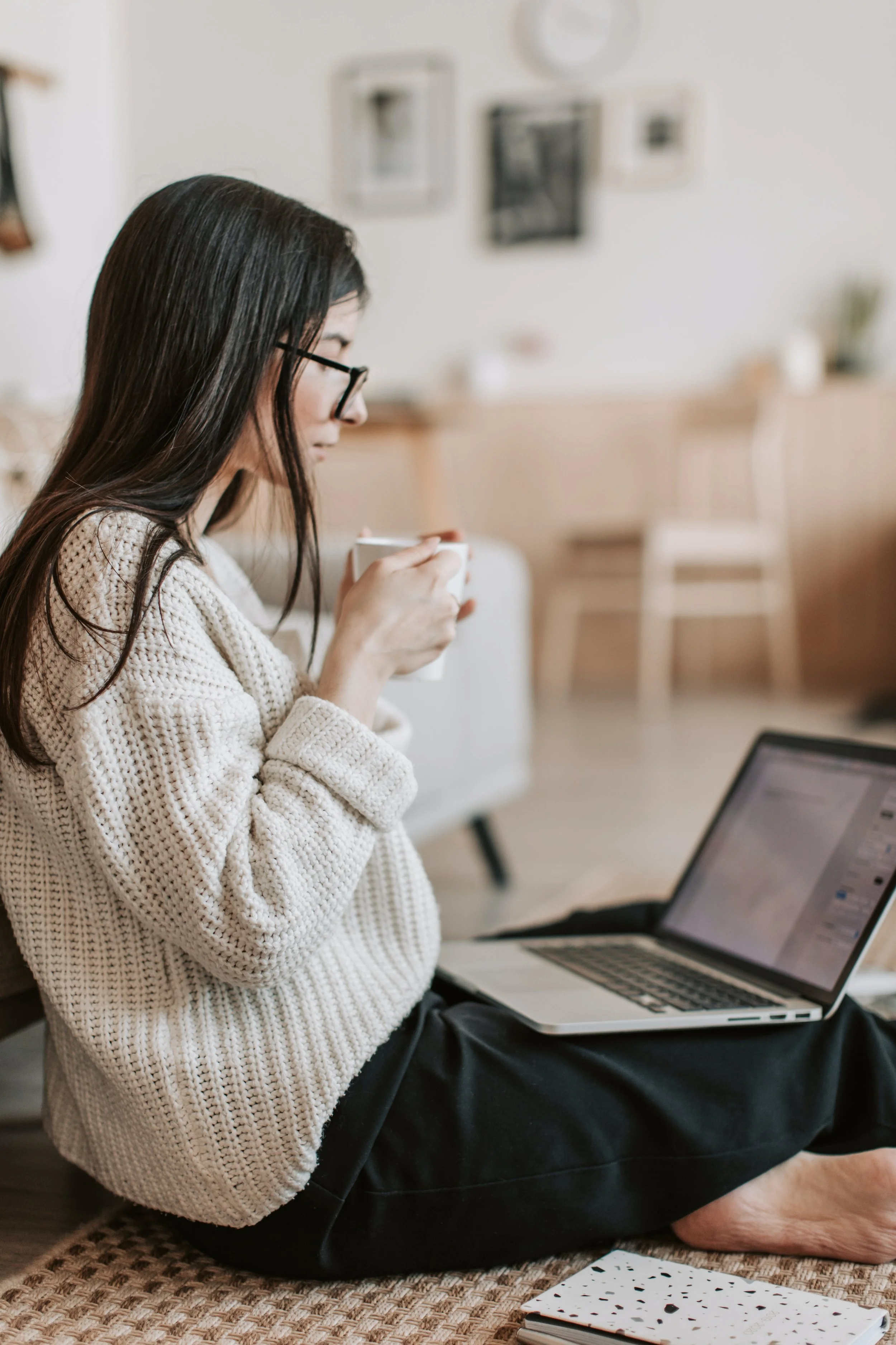 A woman with long dark hair and glasses, wearing a beige knit sweater and black pants, sitting cross-legged on a woven mat with a laptop and holding a white mug, in a cozy, well-lit room with a white wall and framed pictures in the background.