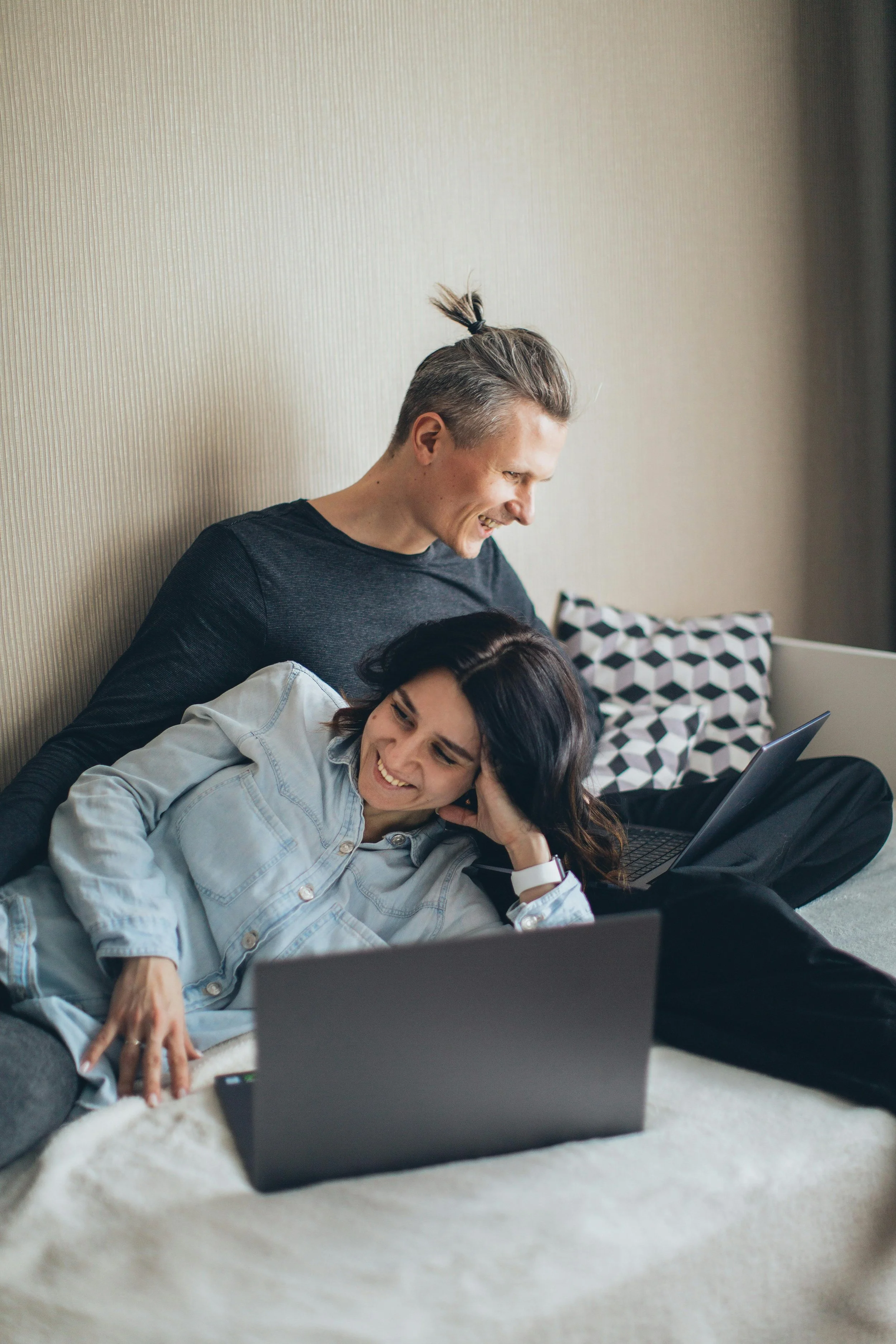 A young couple laughing and relaxing on a couch with laptops in front of them.