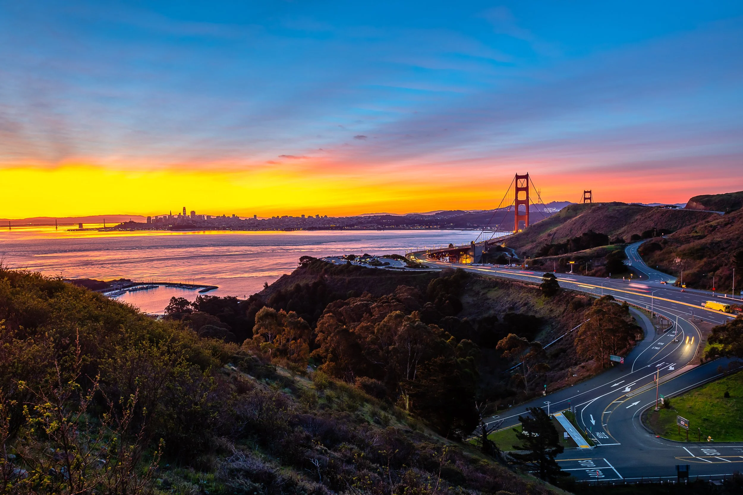 Sunset view of the Golden Gate Bridge in San Francisco, California, with a winding road in the foreground, hills covered with shrubs and trees, and the city skyline in the distance across the water.